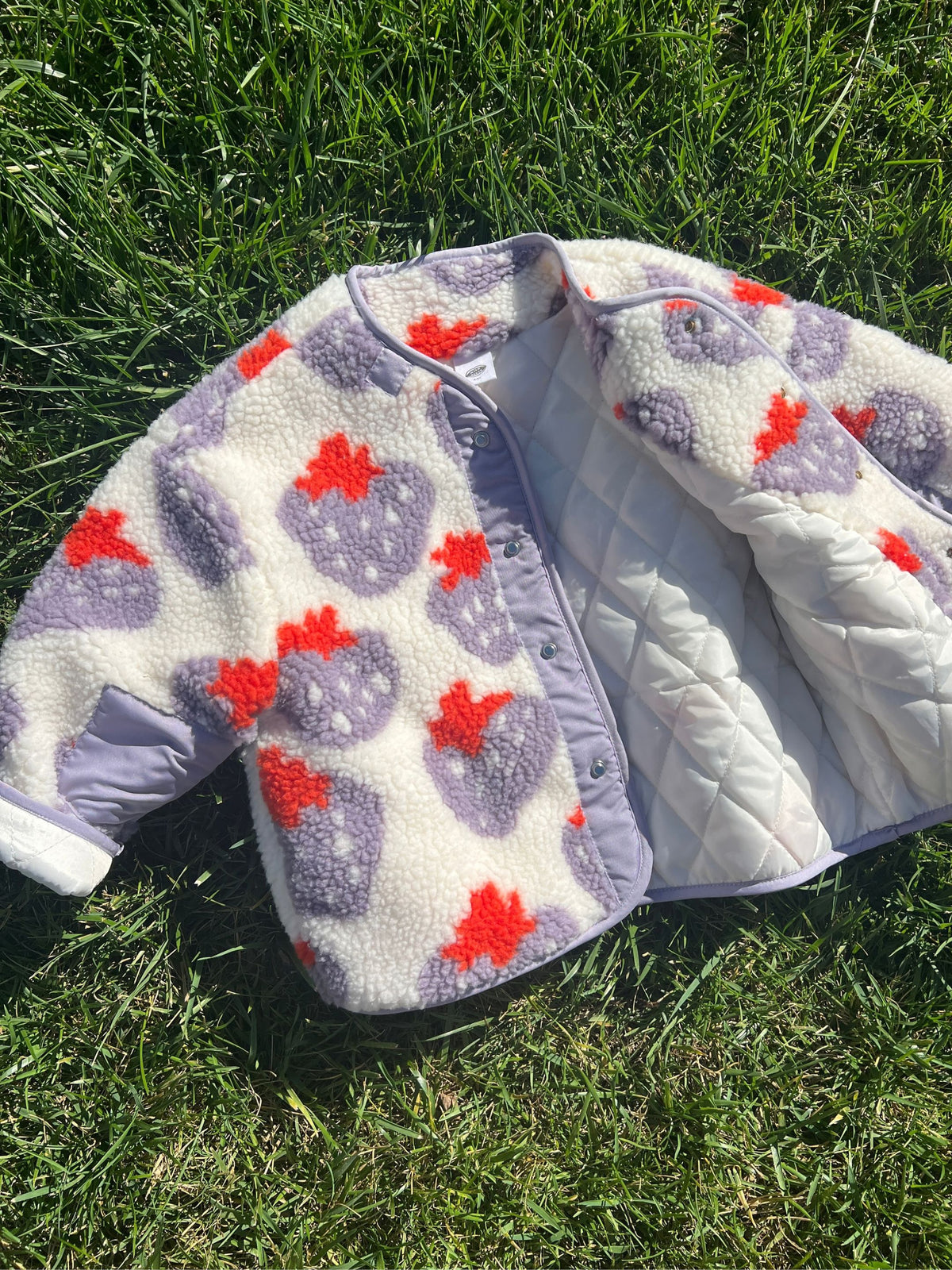 Front view of kids collarless sherpa jacket in cream fleece patterned all over in a purple and red strawberry print, with front snap closure open to show white quilted lining; shown laying on grass