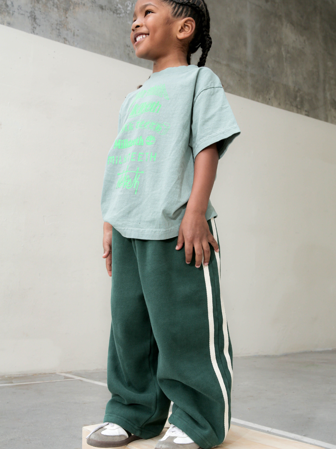 Hunter | Child wearig the varsity track pants in hunter green with white stripes down the side. He wears his hair in braids, a light green t-shirt with neon green Milk Teeth lettering, and white and grey sneakers. He is smiling and standing on a wooden box on a grey concrete playground.