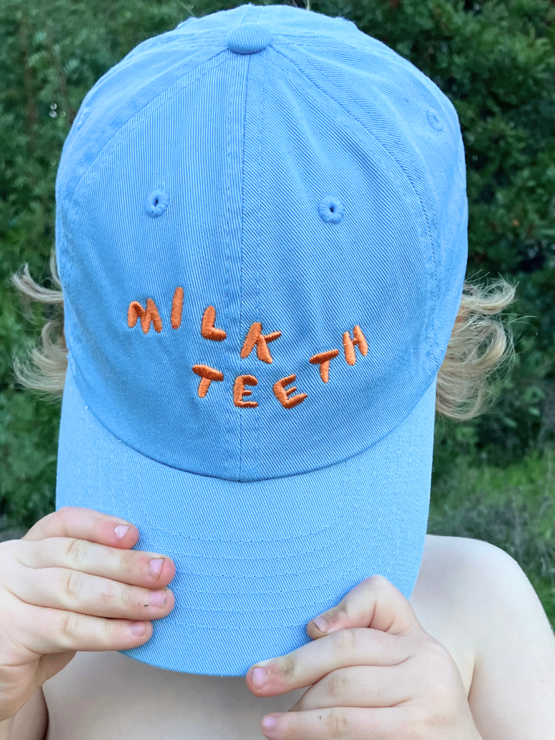 Child wearing the Milk Teeth embroidery caps in blue wit orange lettering, pulling the hat over their face