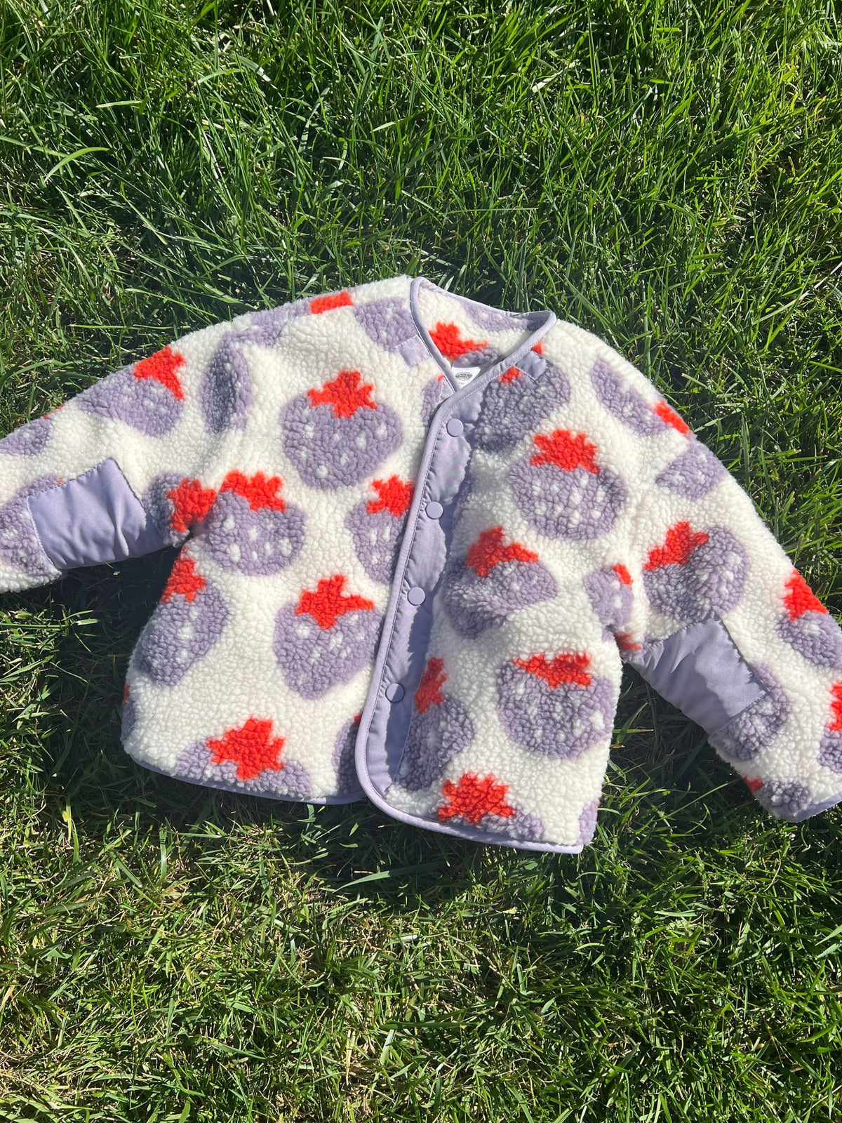 Front view of kids collarless sherpa jacket in cream fleece patterned all over in a purple and red strawberry print, with front snap closure, shown laid flat on grass
