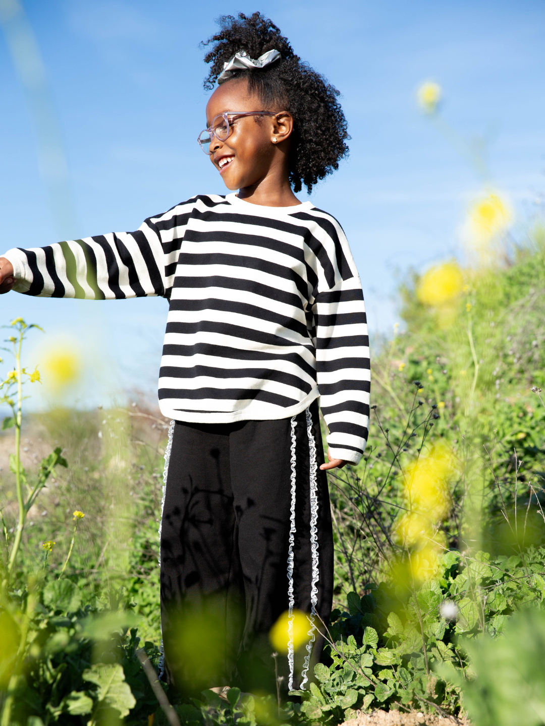 Black | Child wearing the perfect stripey longsleeve tee with black and cream horizontal stripes. She wears black sweatpants with two ruffled stripes down the side, pink glasses, and a silver scrunchie in her curly hair. She is smiling and standing in a green field with yellow flowers and blue sky.