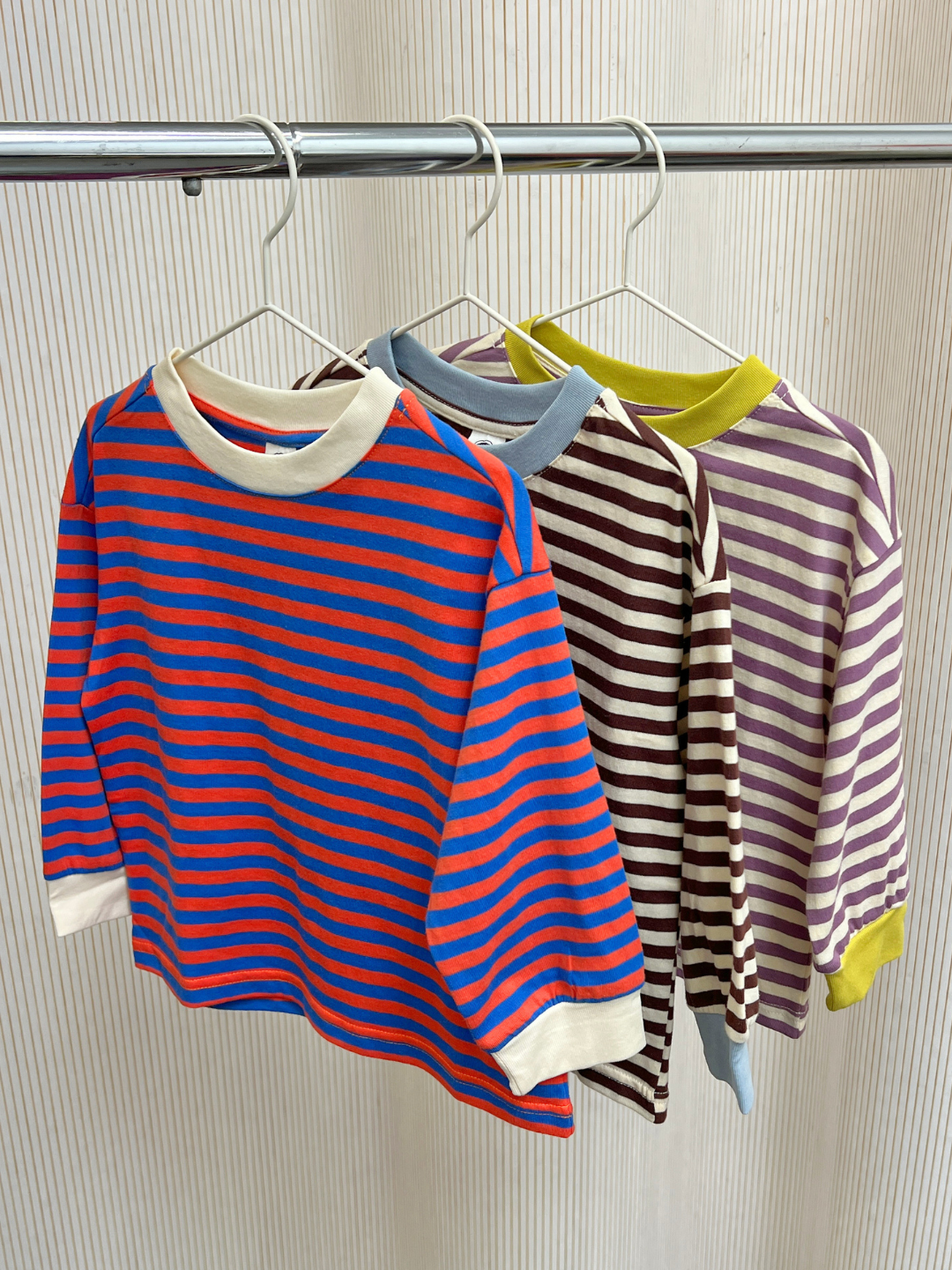 Red/Blue | Three STRIPE LONGSLEEVE shirts, each in distinct red-blue, brown-cream, and purple-cream striped patterns, hang on white hangers from a metal clothing rack. These relaxed-fit long-sleeve tees are made from soft 100% cotton jersey, offering vibrant stripes and effortless everyday style.