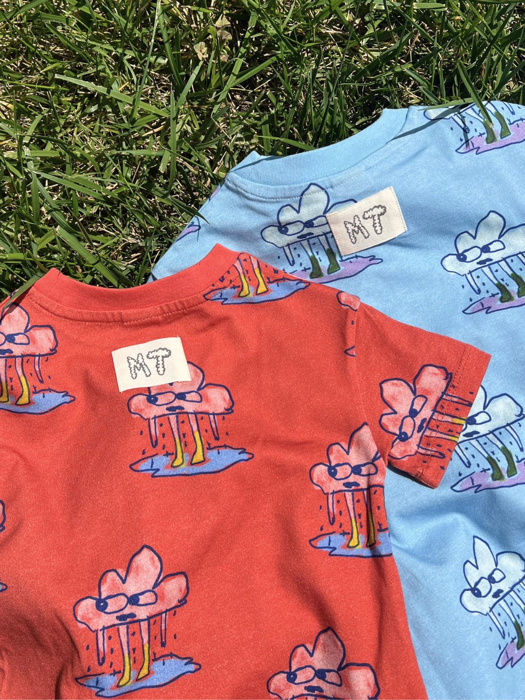Two children's t-shirt rompers, one red and one blue, with cartoon cloud designs on grass.