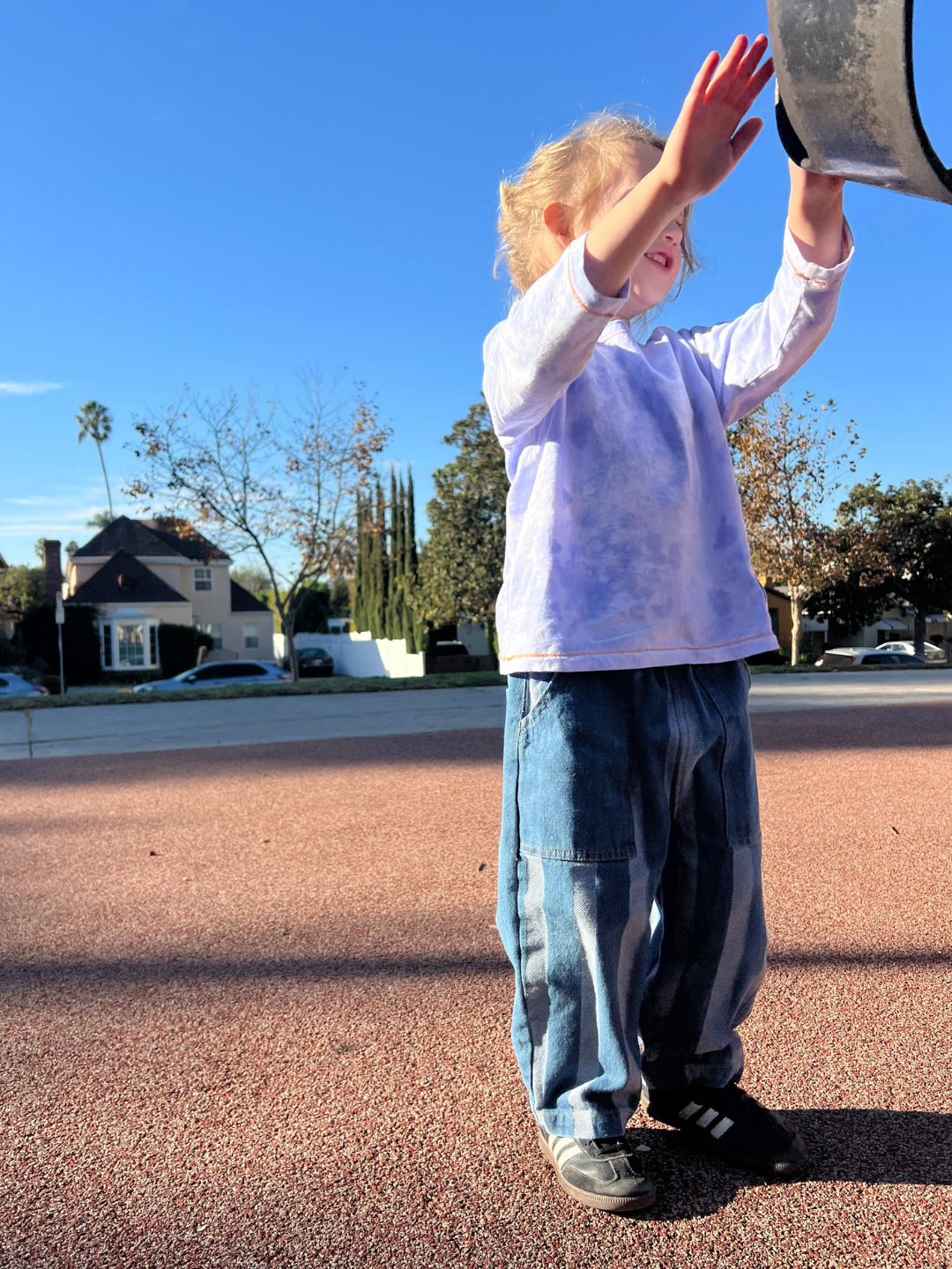 Wearing a KOKO MOMO LONGSLEEVE in a vibrant purple tie-dye, a child joyfully reaches toward the playground equipment on a sunny day, with trees and houses dotting the background. This scene captures vivid imagery, perfectly fitting our product-specific size chart for active kids' wear.