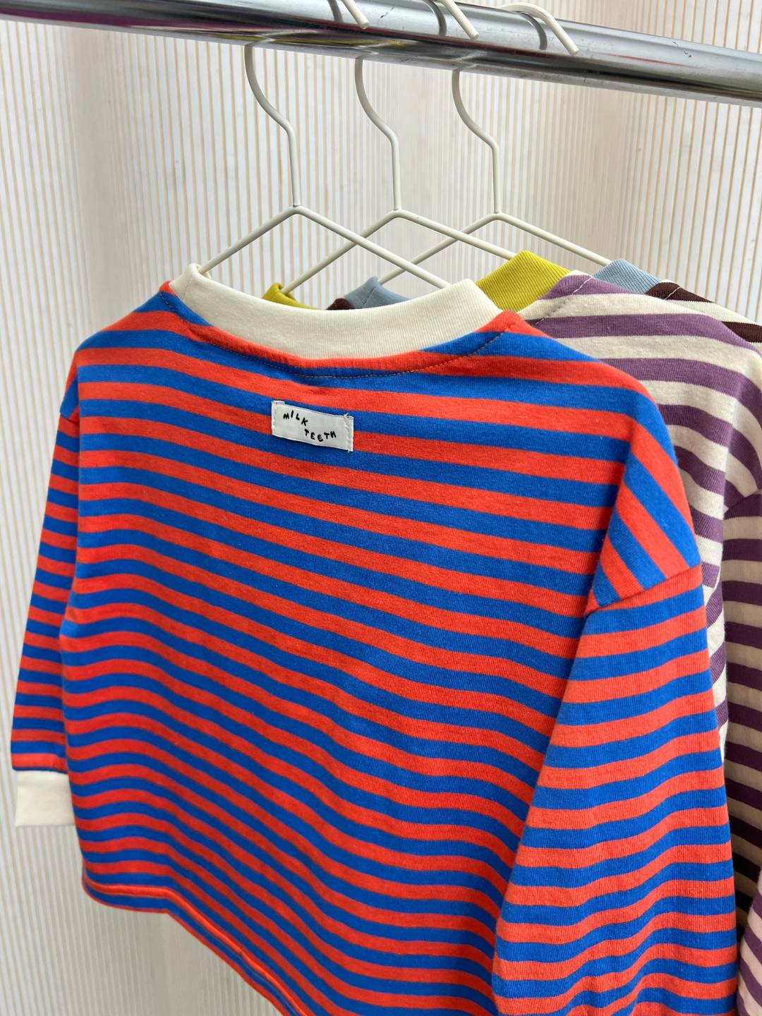 Three STRIPE LONGSLEEVE shirts, featuring blue-red, green-yellow, and purple-beige stripes and crafted from 100% cotton jersey fabric, hang on white hangers on a rack.