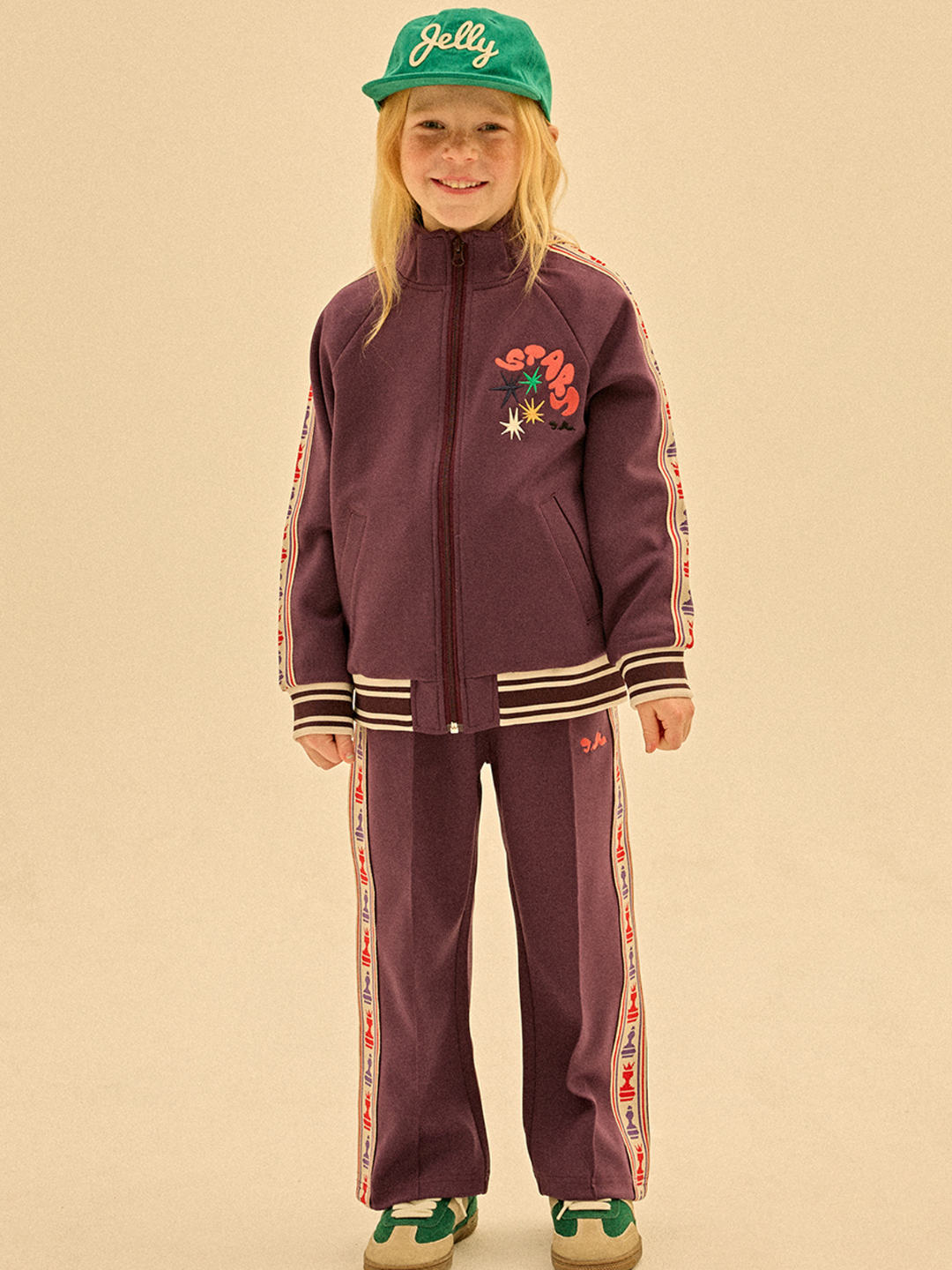 A smiling child wears the CHESS TRACK JACKET in purple with side stripes, paired with a green "Jelly" cap and green sneakers—looking priceless against a plain background