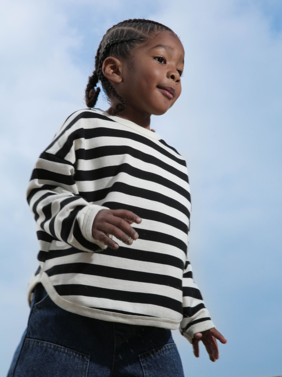 Child wearing the perfect stripey longsleeve tee with black and cream horizontal stripes. He wears blue jeans and his hair in braids, and is standing against blue sky.