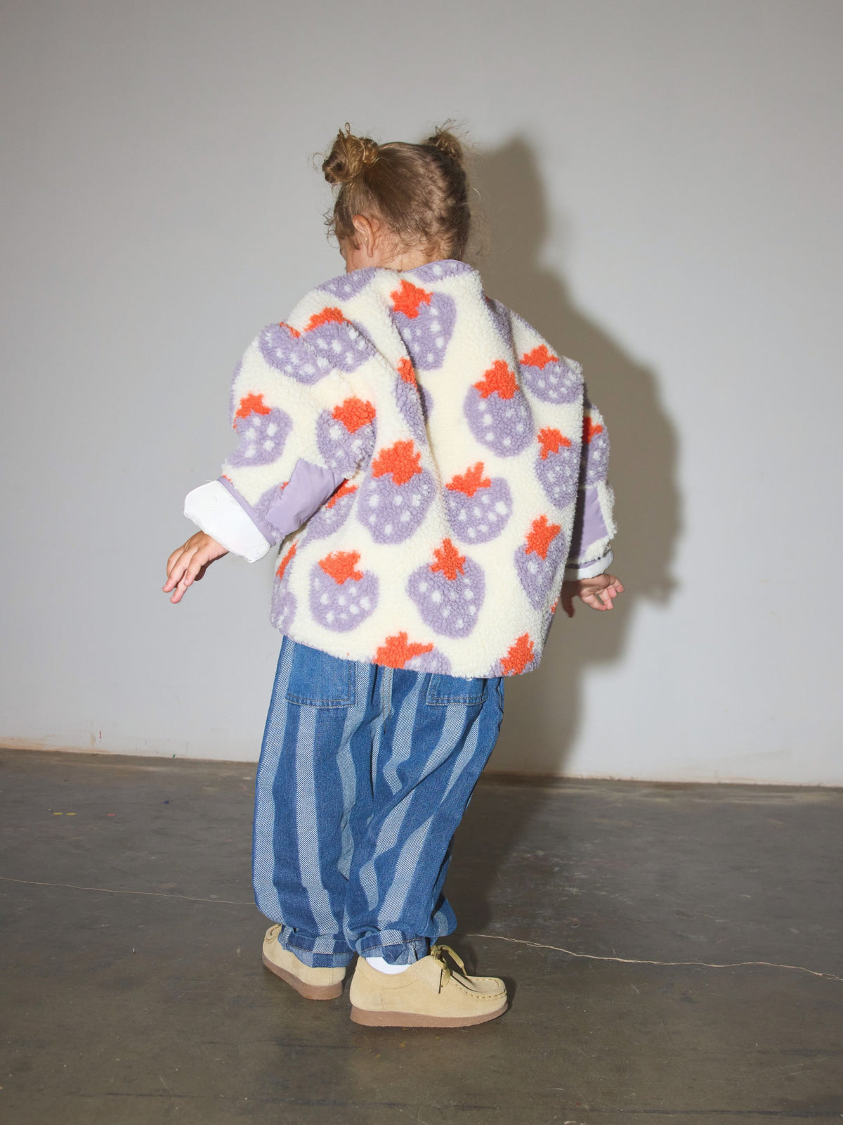 Child wearing a sherpa jacket in cream fleece patterned all over in a purple and red strawberry print, with front snap closure, paired with blue striped jeans and tan shoes, facing away from camera, standing on a concrete floor in front of a white wall