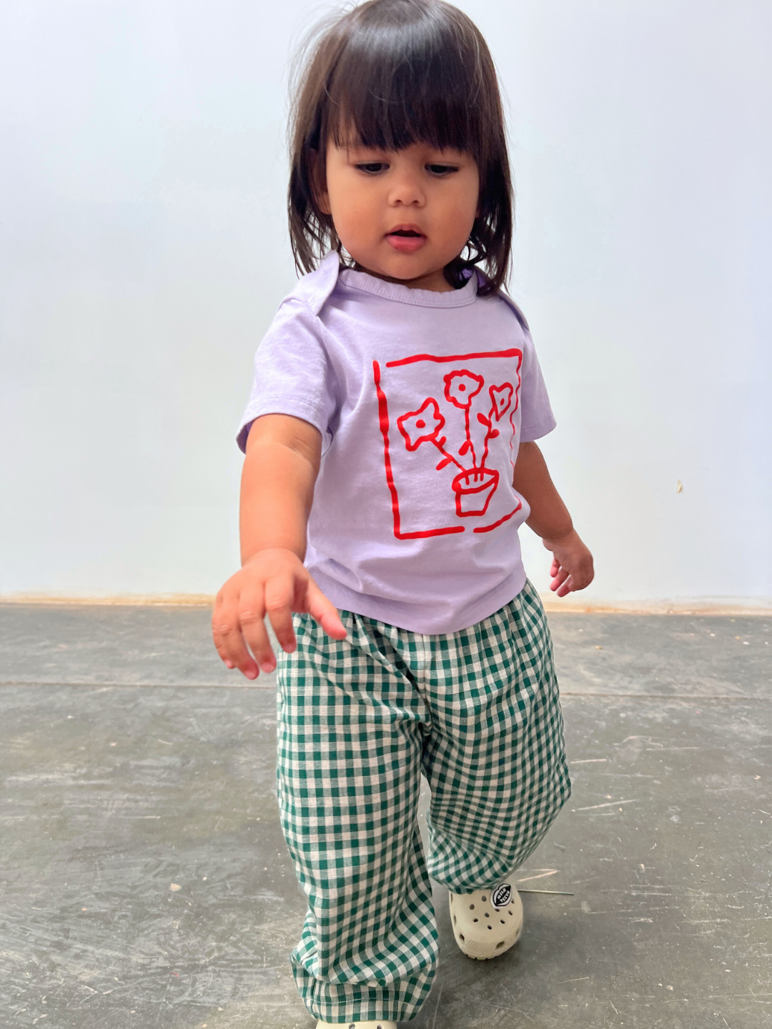 Child wearing a light purple t-shirt with a red graphic and green checkered pants on a plain background