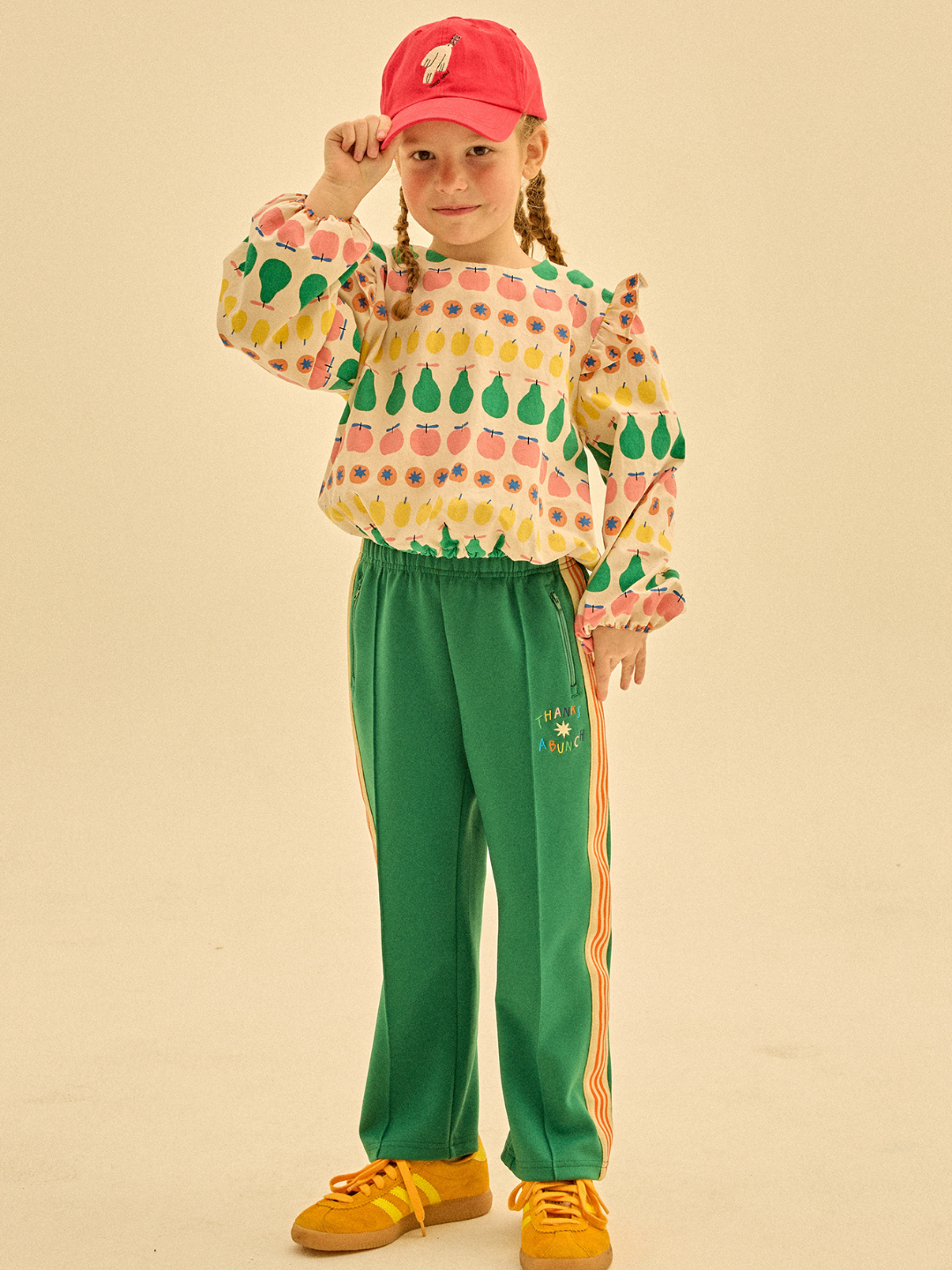 Green | A young girl, dressed in a colorful printed blouse and THANKS TRACK PANTS with side stripes, pairs her outfit with yellow sneakers and a red cap. She stands against a plain background, holding the brim of her cap with one hand to showcase the vibrant ensemble ideal for brand merchandising.