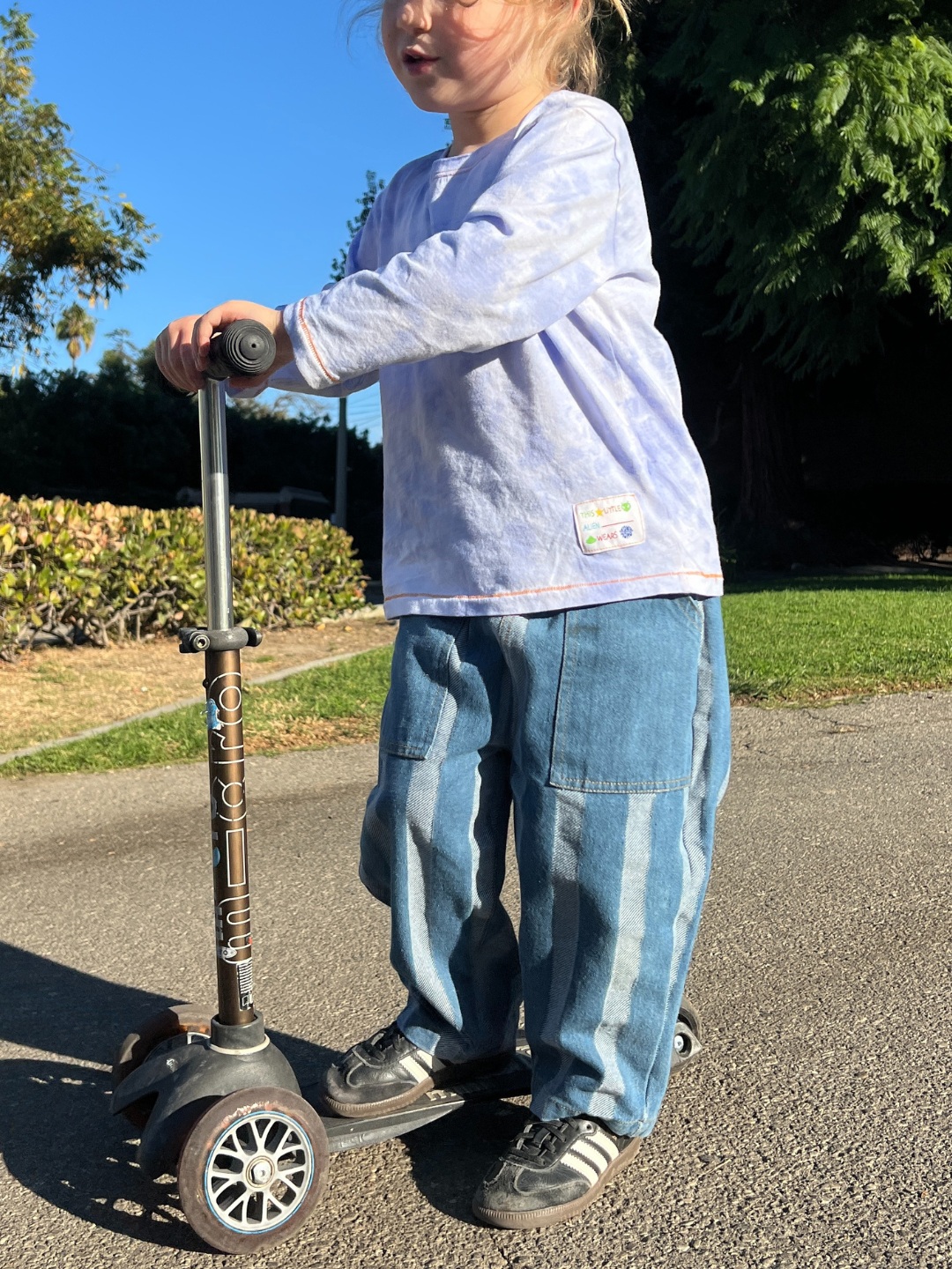 A child wearing the colorful KOKO MOMO LONGSLEEVE and jeans joyously rides a three-wheeled scooter under a sunny sky, creating vibrant imagery. Make your next adventure unforgettable by consulting our product-specific size chart to find the perfect fit.