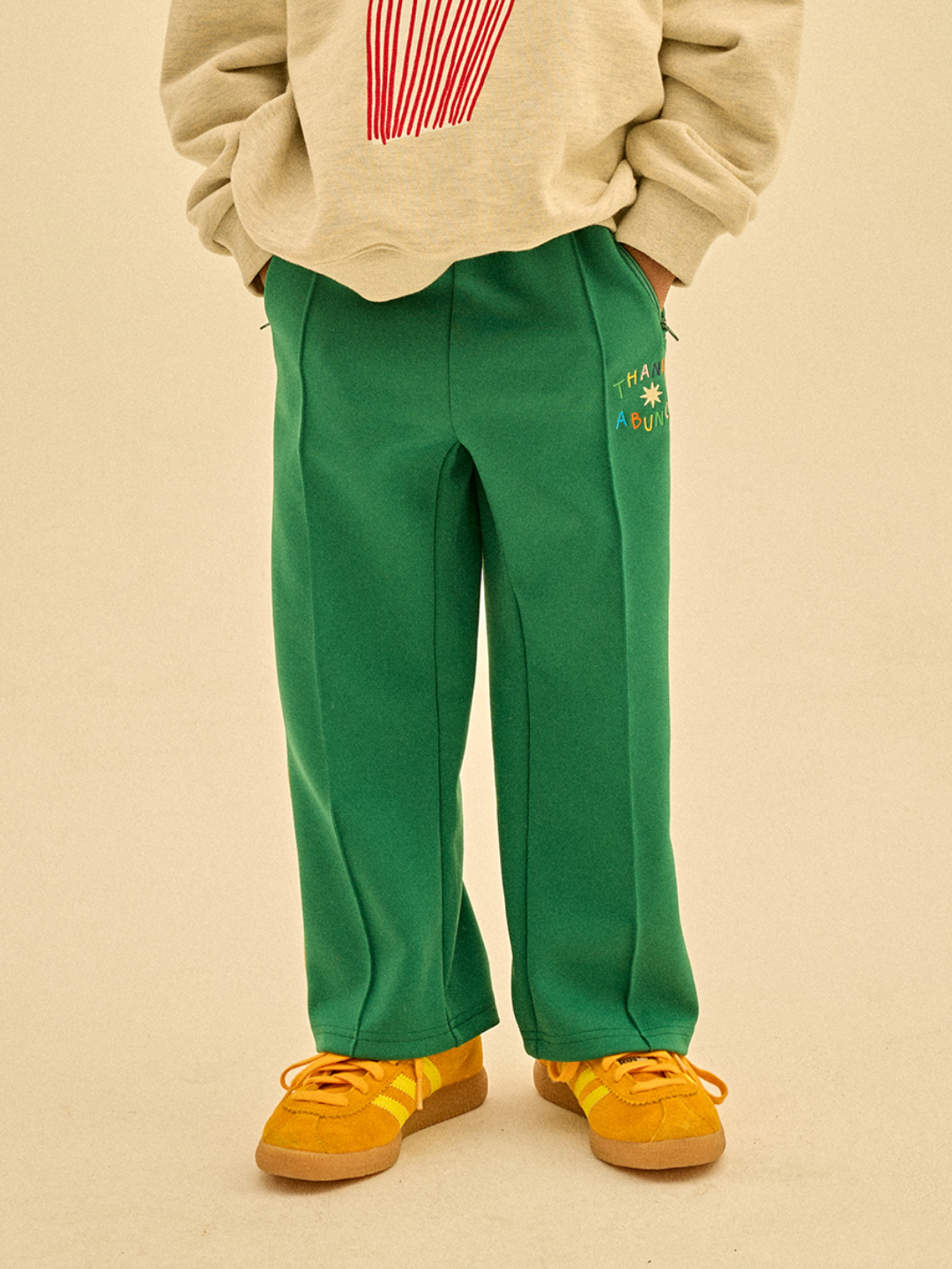 Green | A person wearing THANKS TRACK PANTS with "Thank a Bunch" text, a beige long-sleeve shirt, and yellow shoes stands with their hands in their pockets, showcasing thoughtful merchandising.