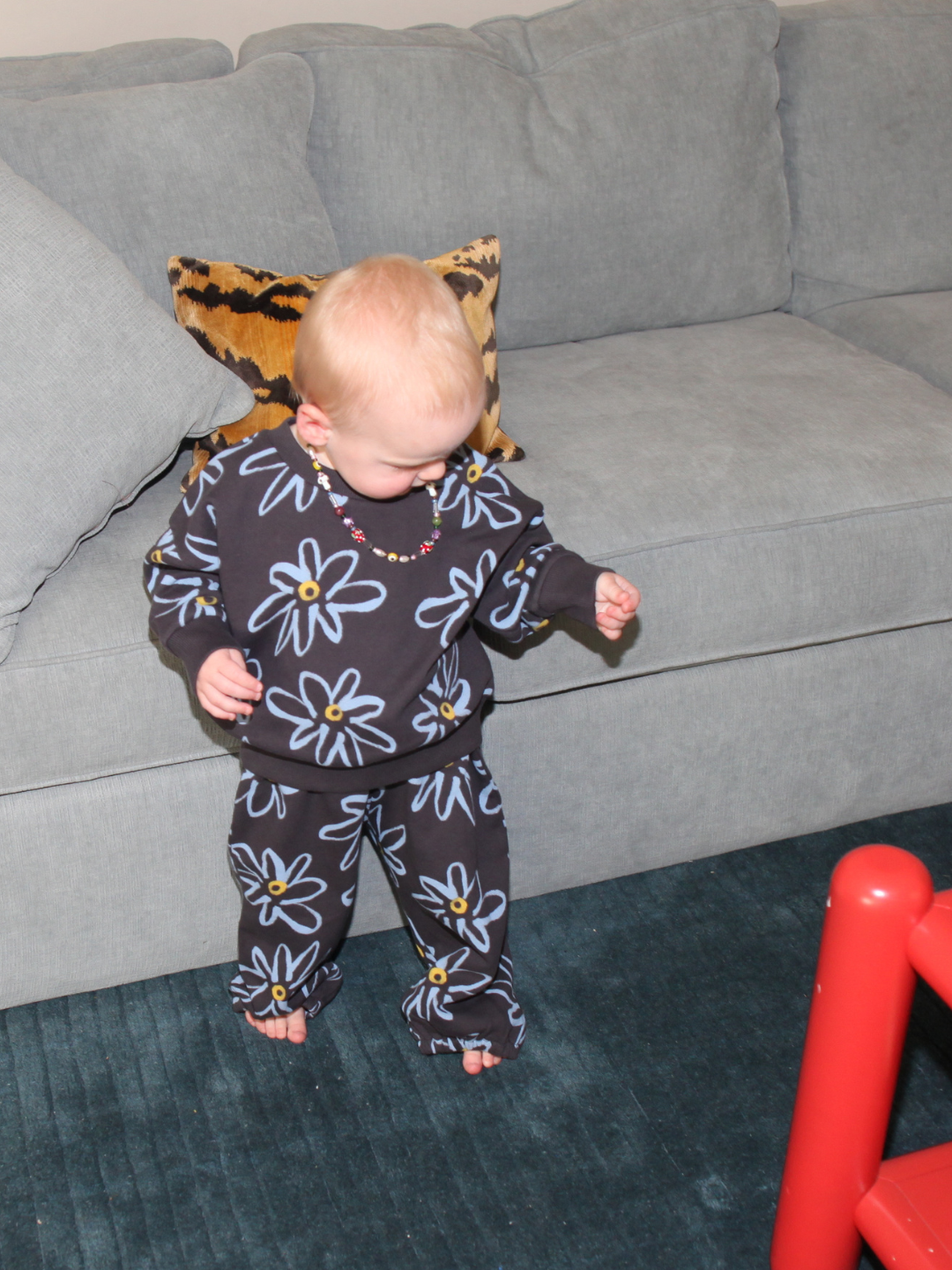 Charcoal | A young child wears the CHARLIE SWEAT SET, made from organic cotton with a painted flower print, standing barefoot on a blue rug in front of a gray couch with a leopard print pillow.