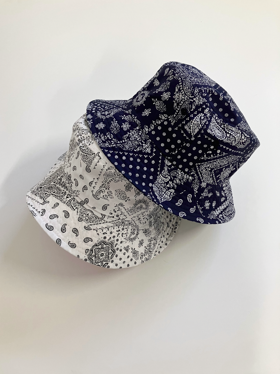 A navy and white bandana bucket hat, one on top of the other on a white surface