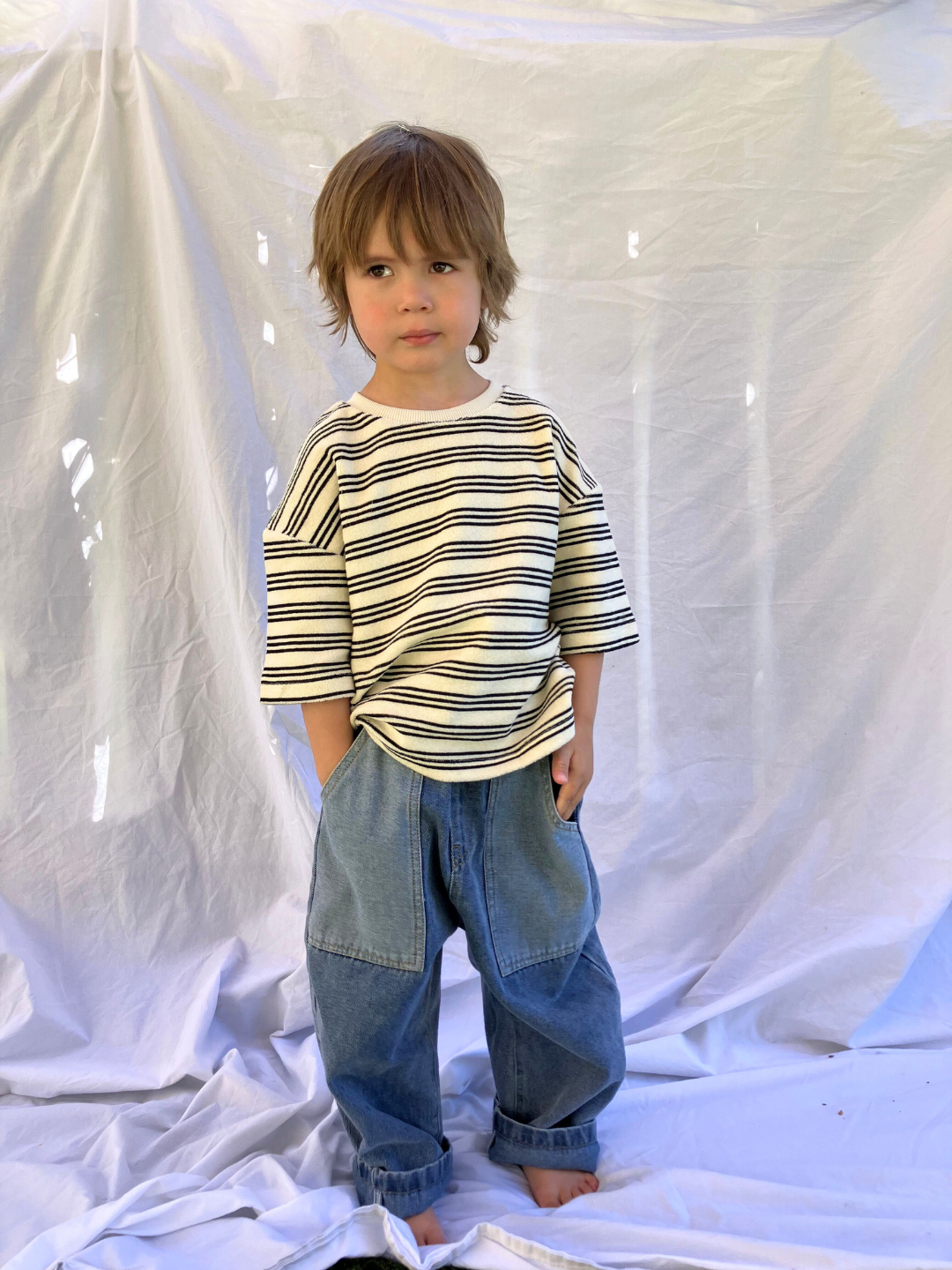 A child wearing the Double Trouble baggy jeans in medium-blue denim with two large patch pockets of lighter denim. He wears an oversized striped t-shirt in cream, with black stripes, and stands in front of a white sheet.