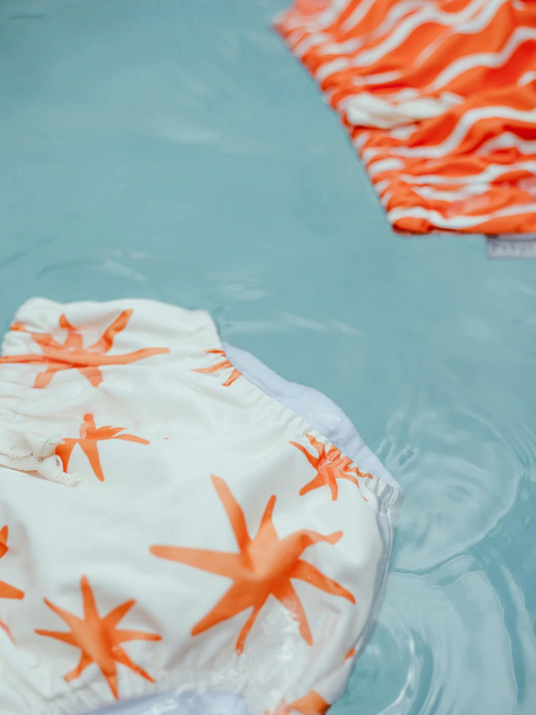 The front view of the swim diaper with an elastic waist with a tie and elastic leg holes. The diaper is a cream color with orange stars shapes all over. It is floating in a pool with the Spaghetti Swim Diaper.