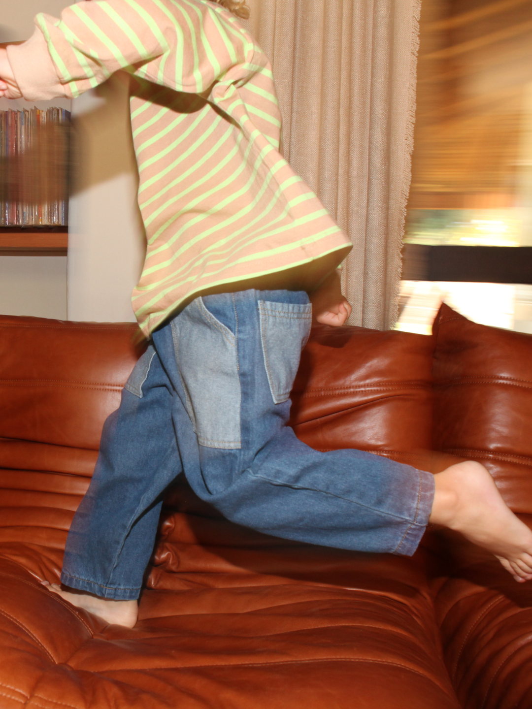A child wearing a striped shirt and baggy denim pants jumping on a brown leather couch.