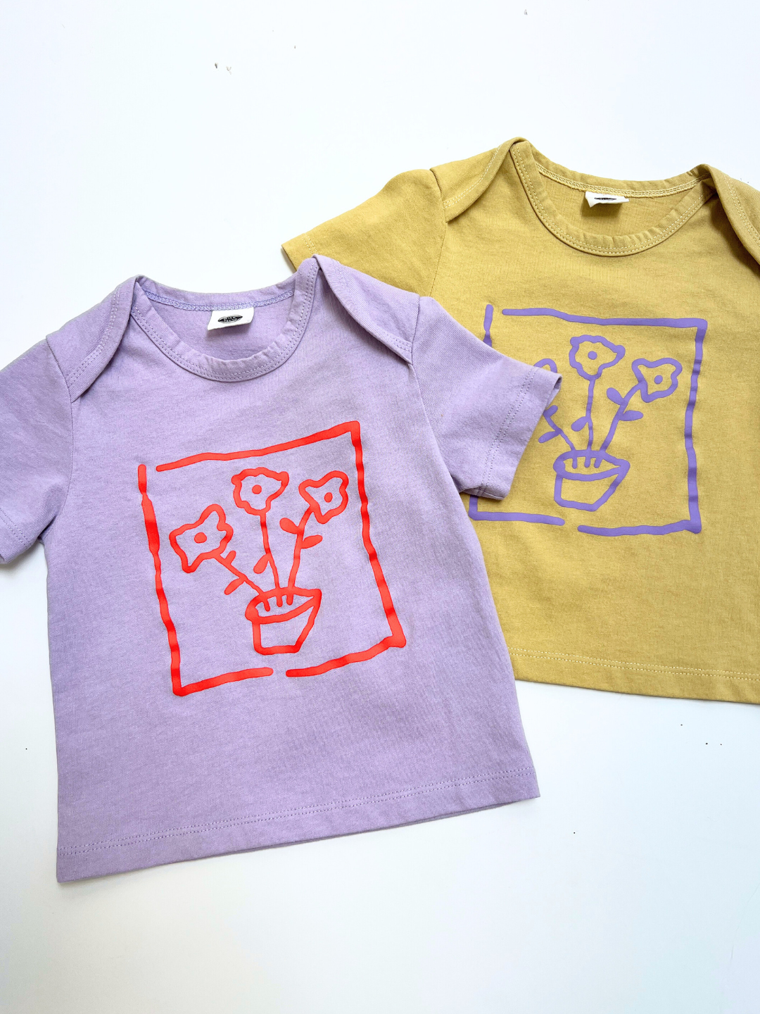 Two FLOWER POT BABY TEE shirts for kids—one in lavender with a red flower pot graphic, the other in mustard yellow with a purple flower pot graphic—are displayed side by side on a white surface.
