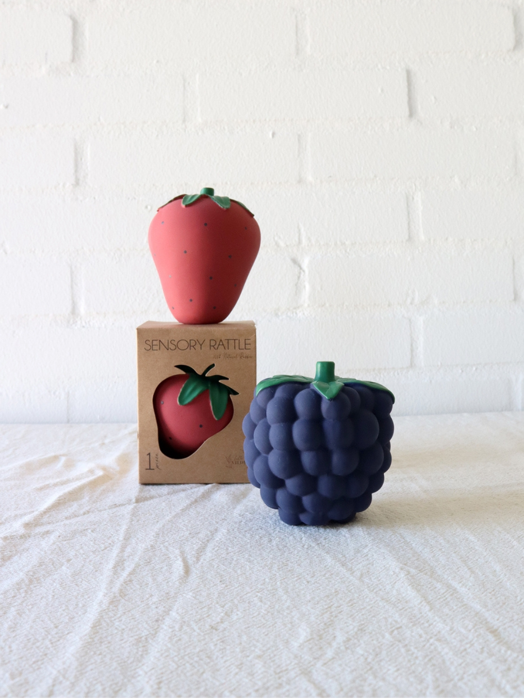 Two BERRY RATTLE TEETHER toys, handcrafted from natural rubber and shaped like a strawberry and a blackberry, are displayed on a white surface. The strawberry rattle is still in its packaging, while the blackberry rattle is positioned in front, ideal for soothing sore gums or serving as a sensory toy.