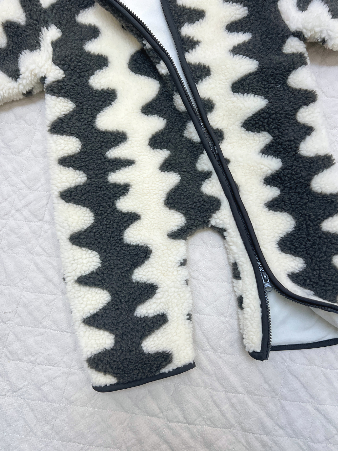 The WAVELENGTH SHERPA FLEECE STROLLER SUIT, featuring a fuzzy black and white zigzag pattern and convenient zipper, is perfect as a cozy winter layer and is shown lying on a quilted white surface.