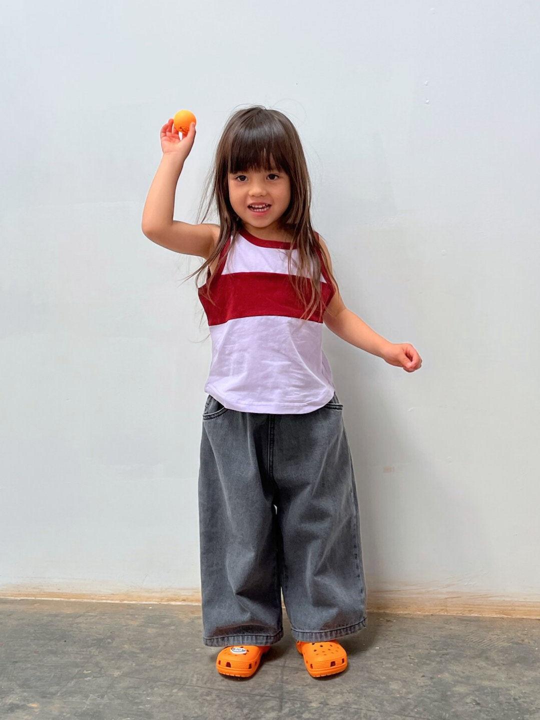 A young child stands indoors by a plain wall, wearing the TRACK TANK, relaxed fit wide-leg jeans, and orange clogs, raising one arm while holding an orange ball.