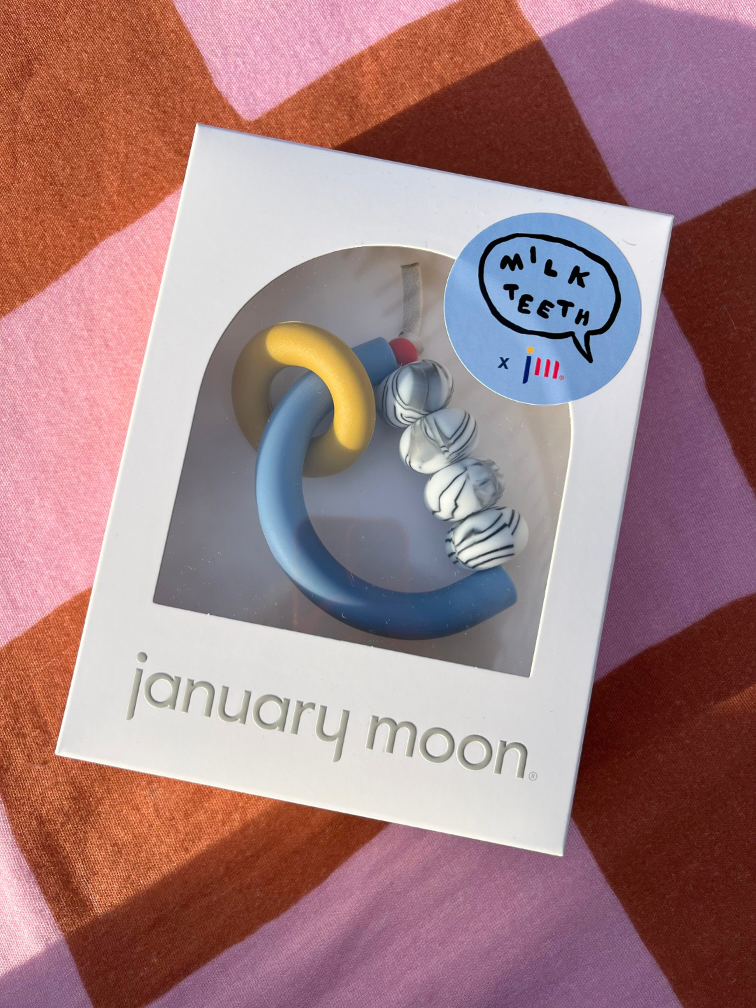 Memphis | Box with MT X JM ARCH RING TEETHER, featuring blue and yellow marbled beads, labeled “january moon,” rests on a pink-orange checkered fabric—a sensory-friendly teether made in the USA.