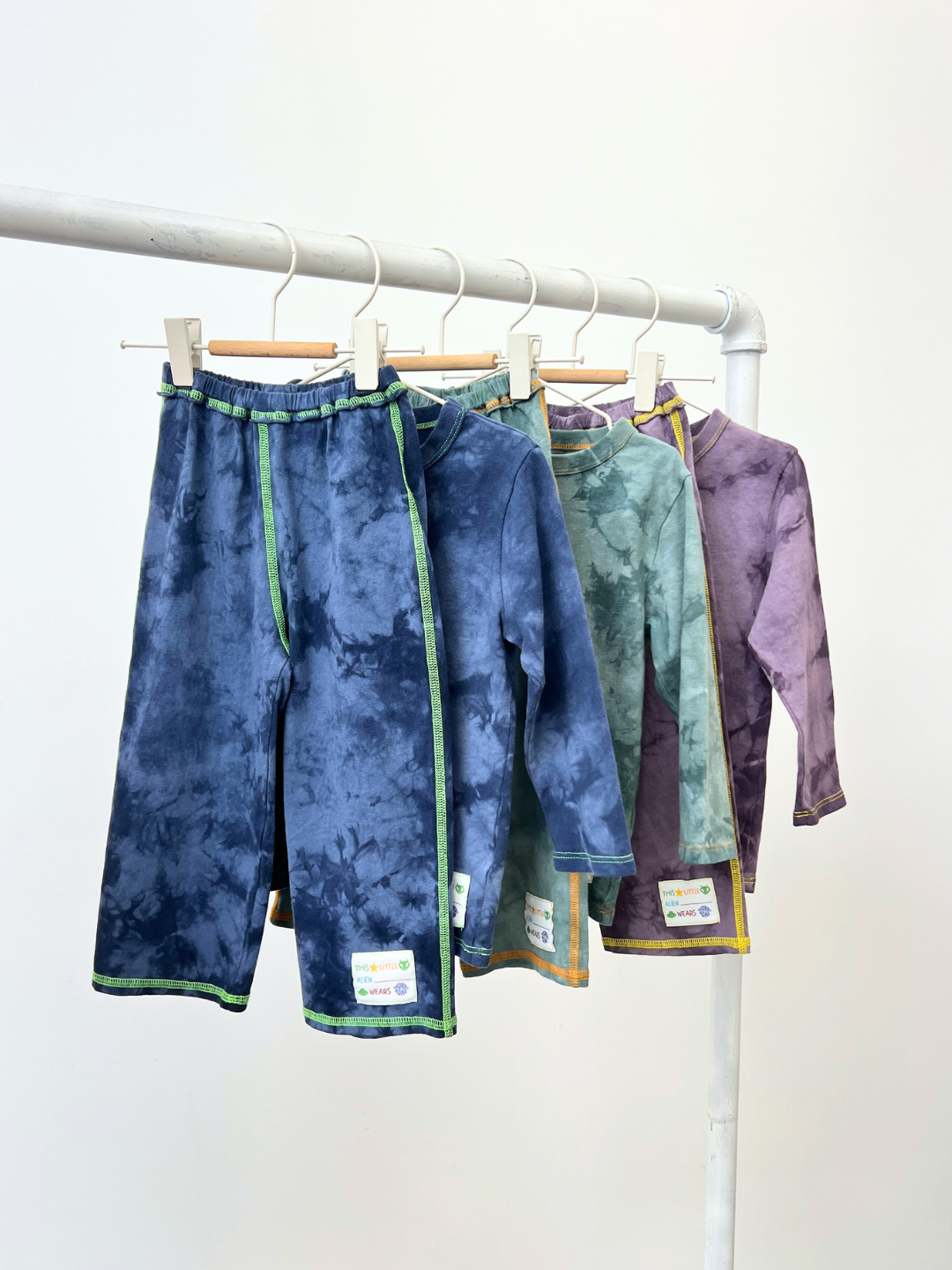 Four sets of COSMO TIE DYE PANT and matching children’s tie-dye long-sleeve shirts in blue, green, and purple hang on a white clothing rack.