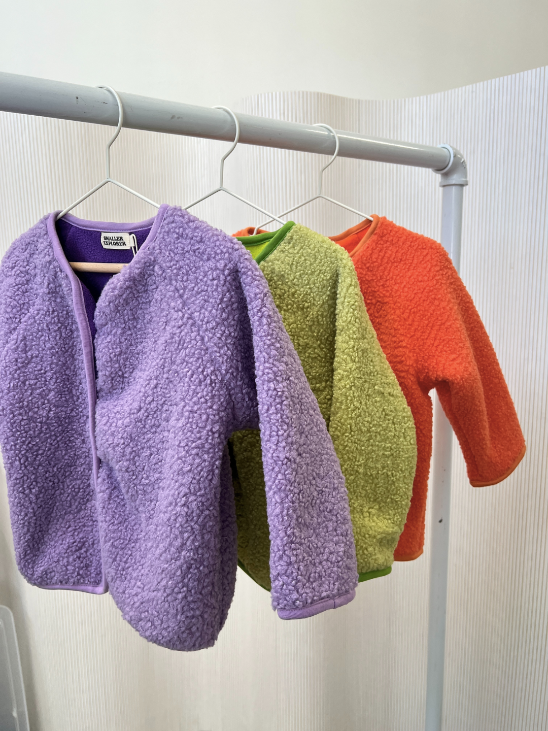 Three kids collarless fleece jackets hand on a white garment rack, on white hangers, in a white room. The jackets are purple, light green, and orange.