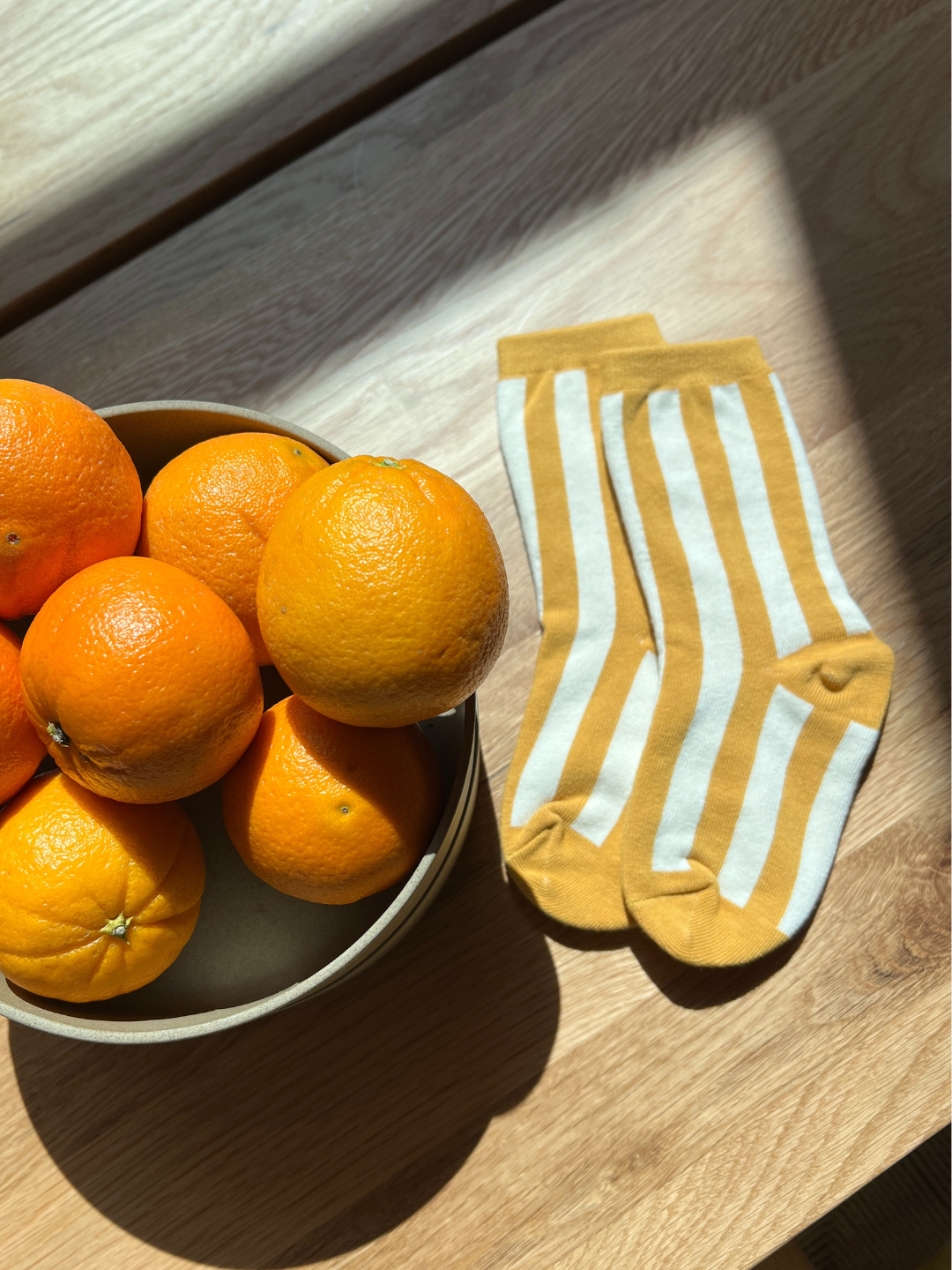 A bowl of oranges sits on a wooden surface next to a pair of comfy, yellow and white STRIPE SOCKs.