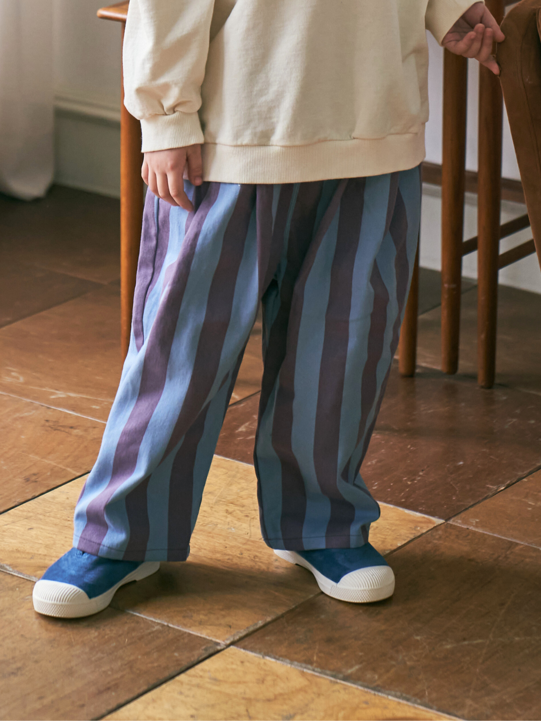 Purple | Cropped waist down image of a child wearing baggy pants in blue with wide purple vertical stripes, standing on a wood tiled floor.