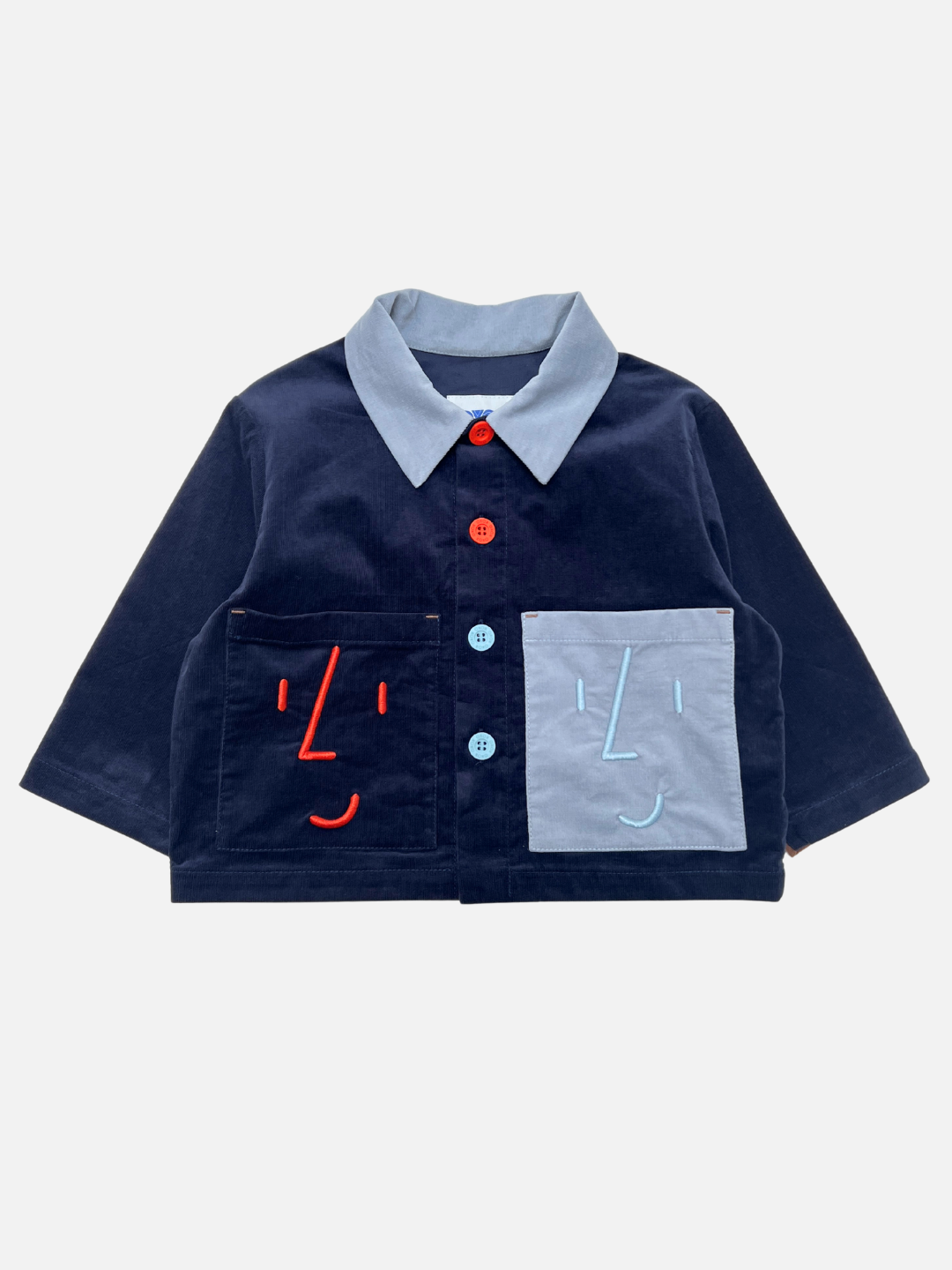 Navy Blue | The FACE JACKET is a navy blue kids corduroy jacket with a light blue collar, mismatched buttons, and two large front pockets embroidered with red and light blue smiling faces.