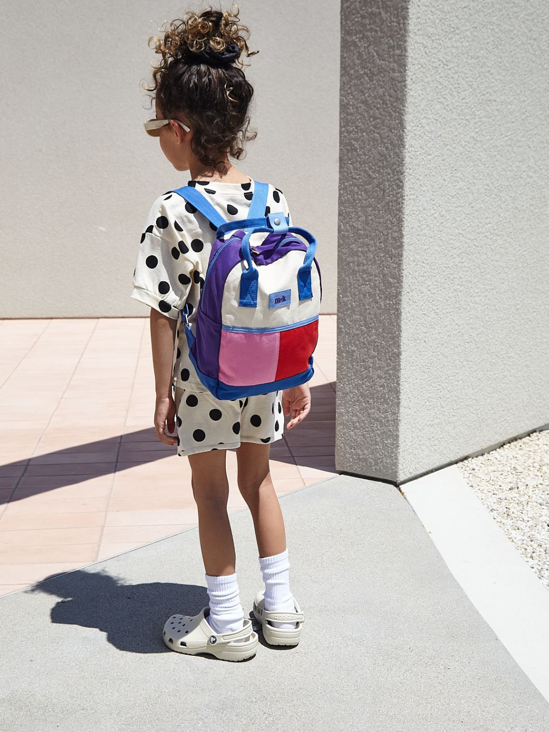 A child wearing a colorblock backpack with bright blue straps, handles and base, purple sides, and one pink and one red patch under a cream top