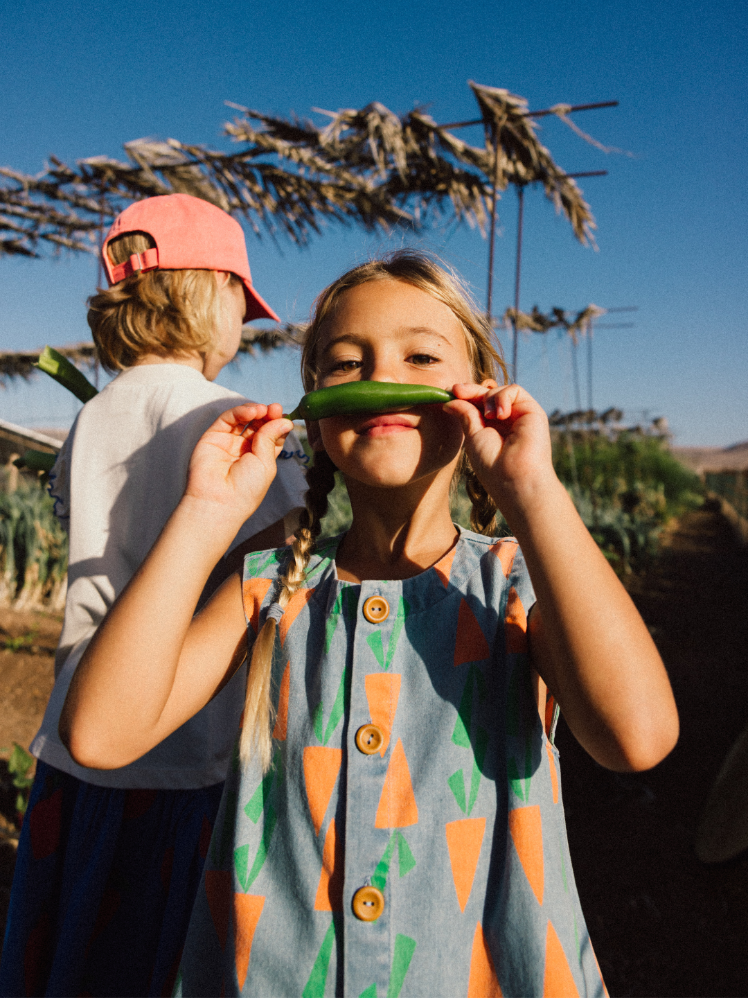 A child with braided hair holds a green vegetable to their face, playfully smiling, while another child in a pink hat and CARROT DENIM VEST stands nearby in a garden under a blue sky.