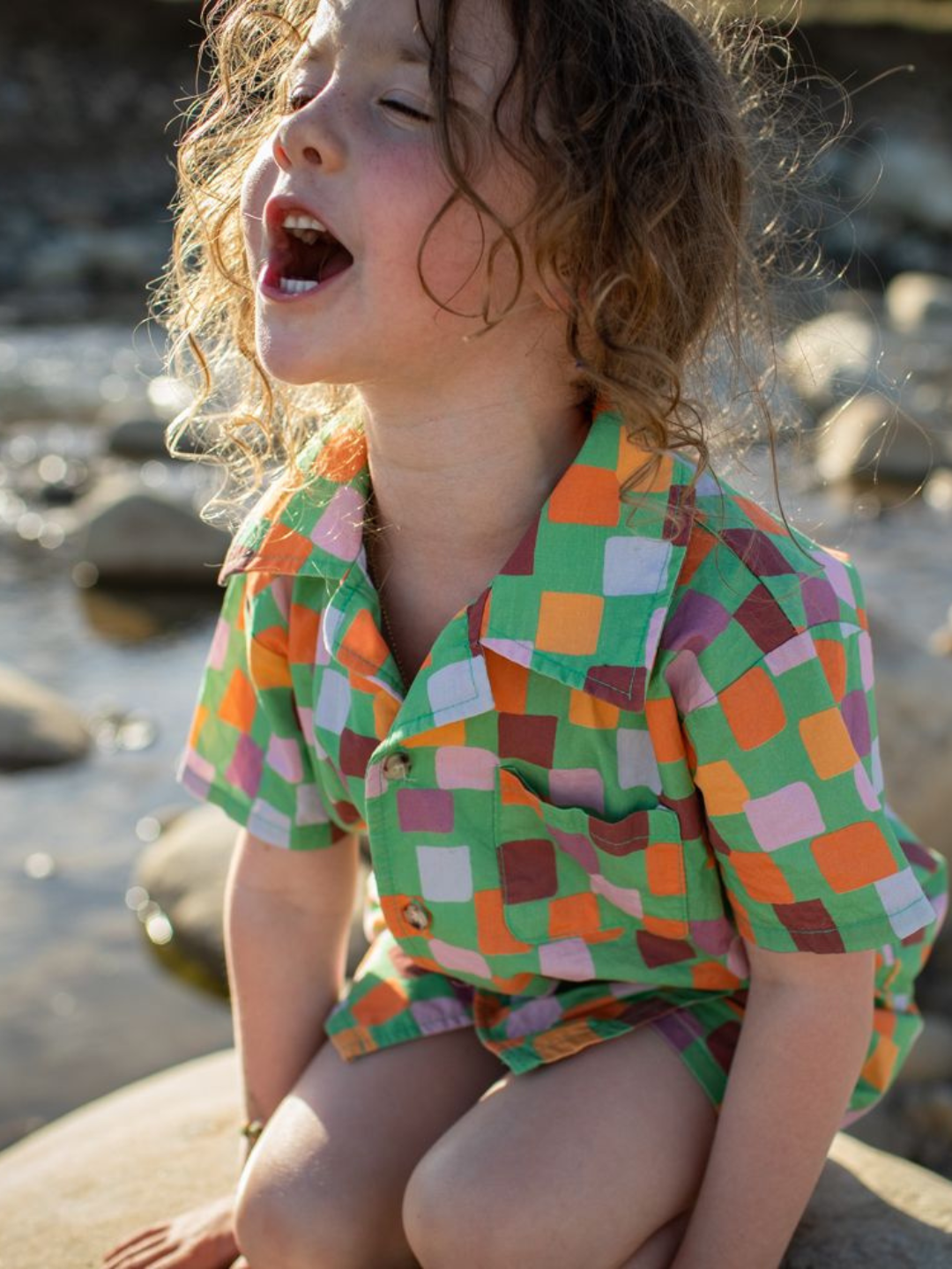 A child sitting on rocks, wearing a kids' shirt and shorts set in a pattern of purple, pink, gold and orange squares on a green background