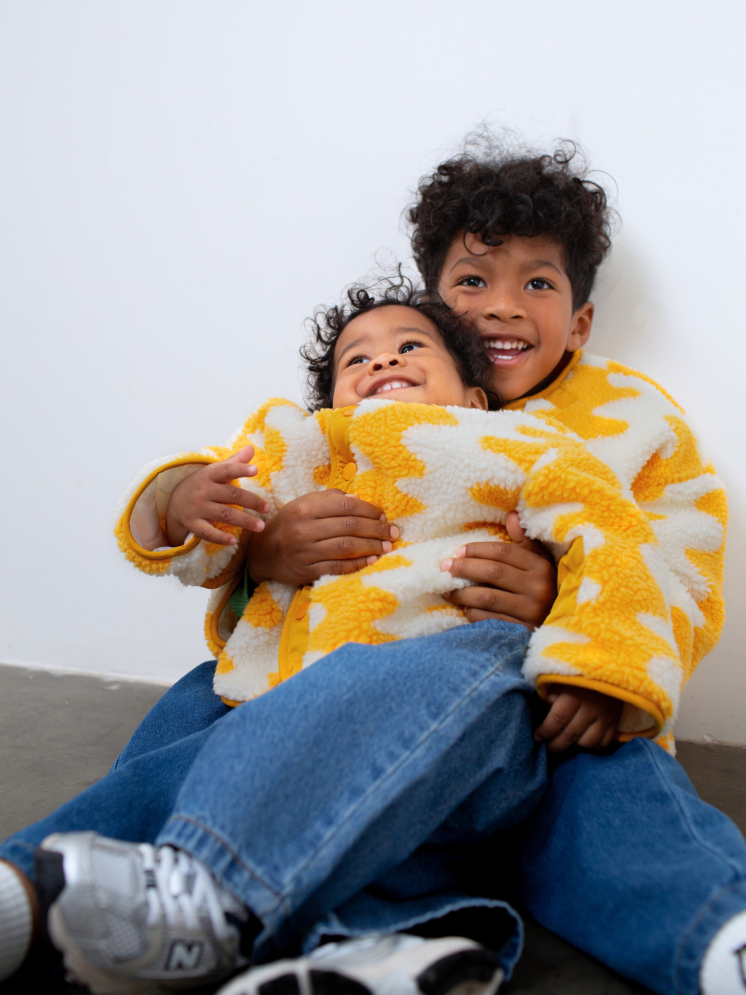 Two children hugging each other wearing yellow and white patterned jackets against a plain background