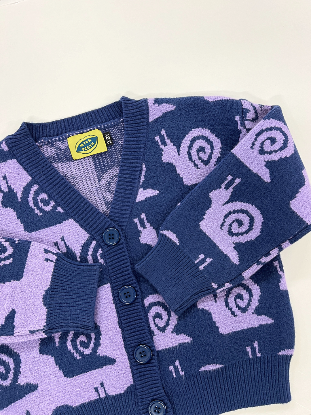 The ESCARGOT CARDIGAN is a navy blue and lavender button-up cardigan adorned with a whimsical snail pattern design. It features three blue buttons and a single pocket on the left side, making it a charming addition that pairs well with both casual and smart attire.