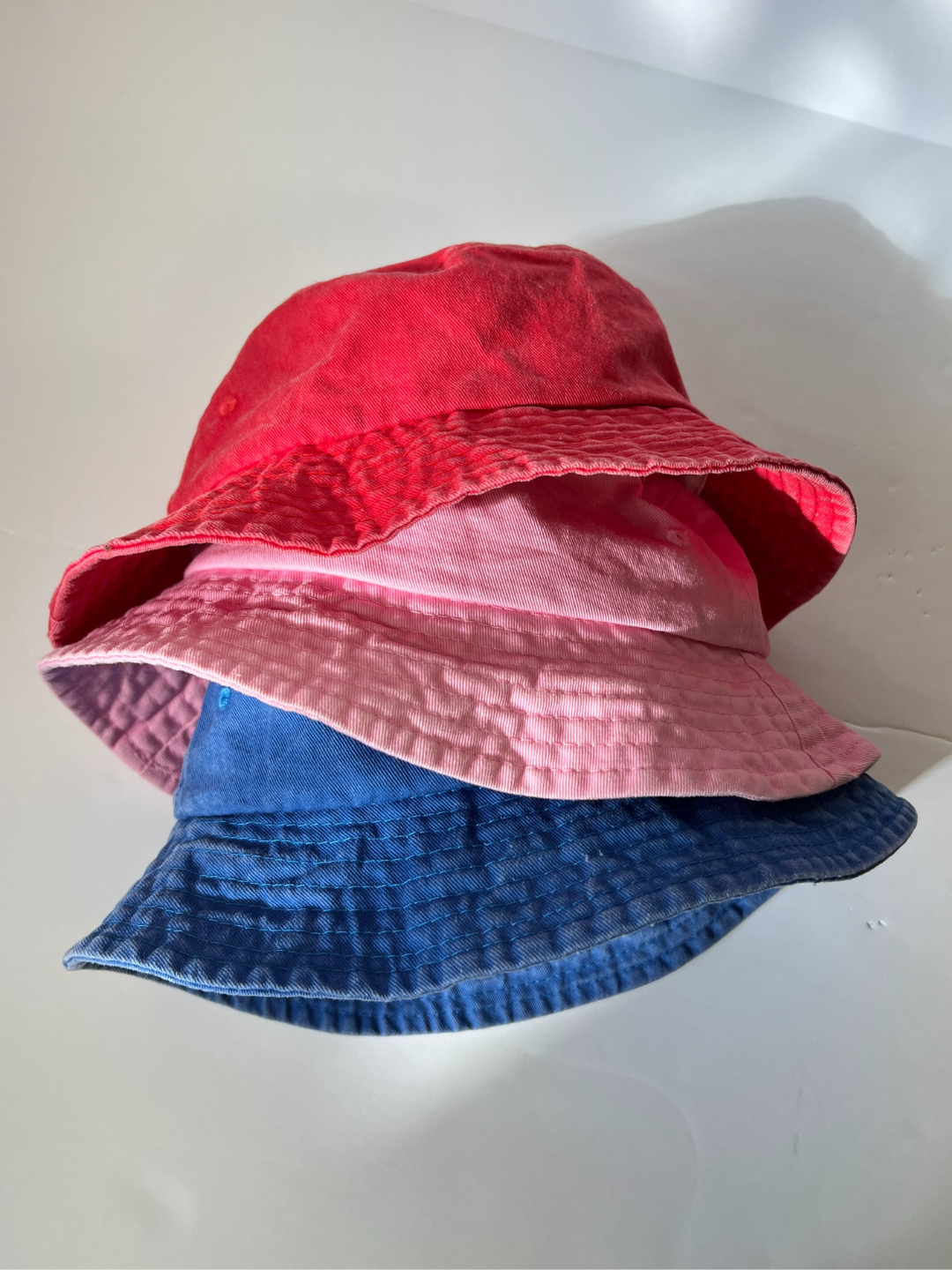 Pink | The PIGMENT BUCKET HAT in red, pink, and blue forms a charming display on a white surface, showcasing a subtle vintage effect that enhances their timeless appeal.