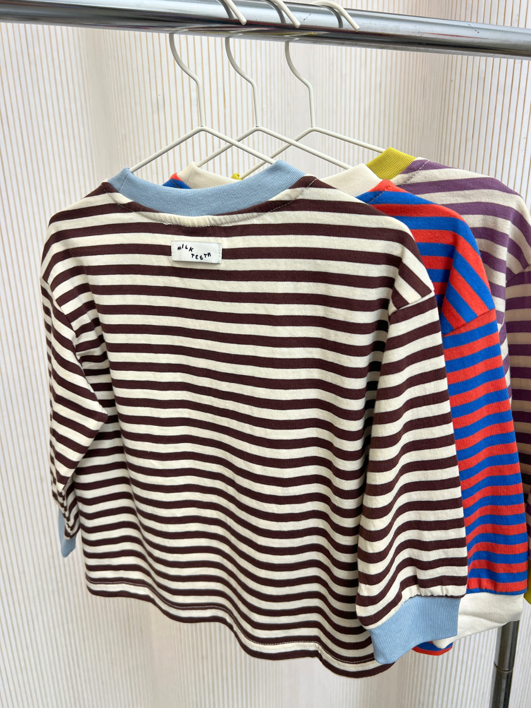 Three STRIPE LONGSLEEVE shirts in various color patterns hang on white hangers from a metal rack against a subtly striped background, each displaying a relaxed-fit silhouette crafted from soft, 100% cotton jersey striped fabric.
