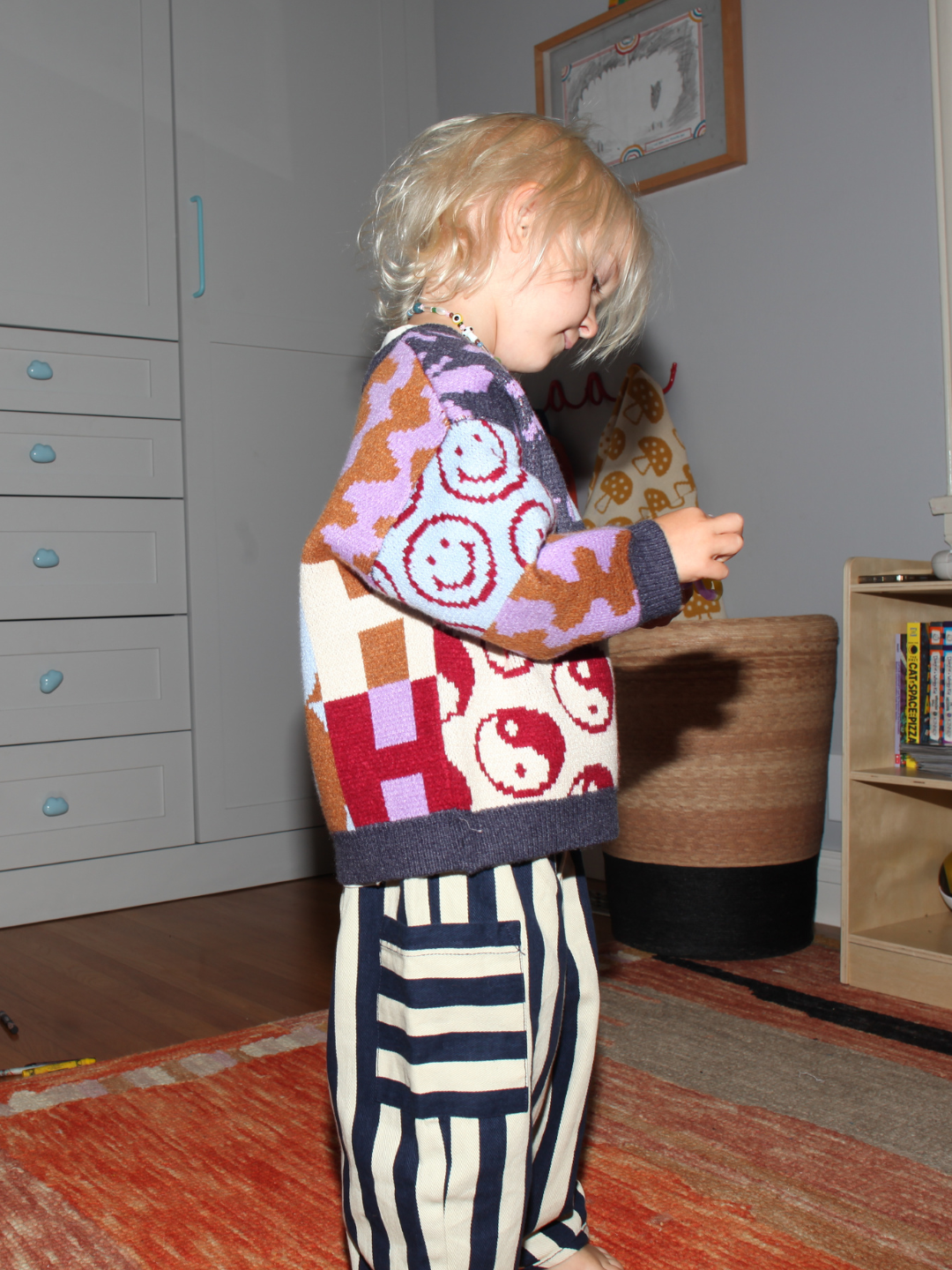 Child wearing a colorful patterned sweater and striped pants in a room with furniture and decor.