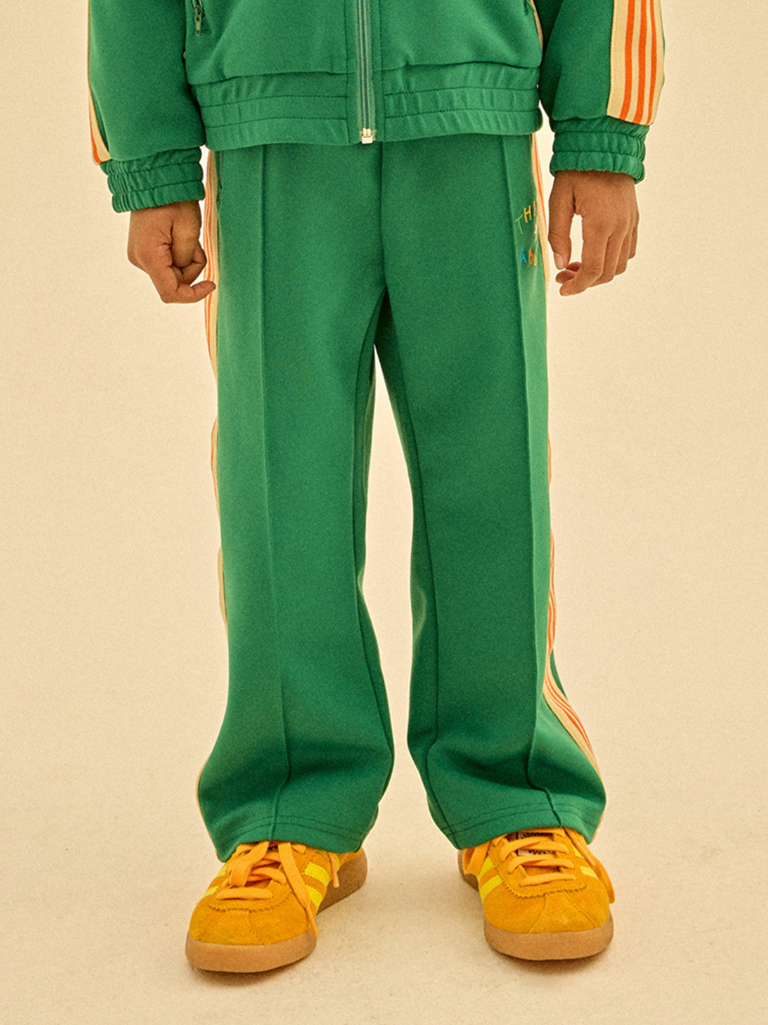 Green | Child wearing the THANKS TRACK PANTS, featuring a green tracksuit with orange stripes and yellow sneakers, stands on a beige background, perfect for your next merchandising campaign.