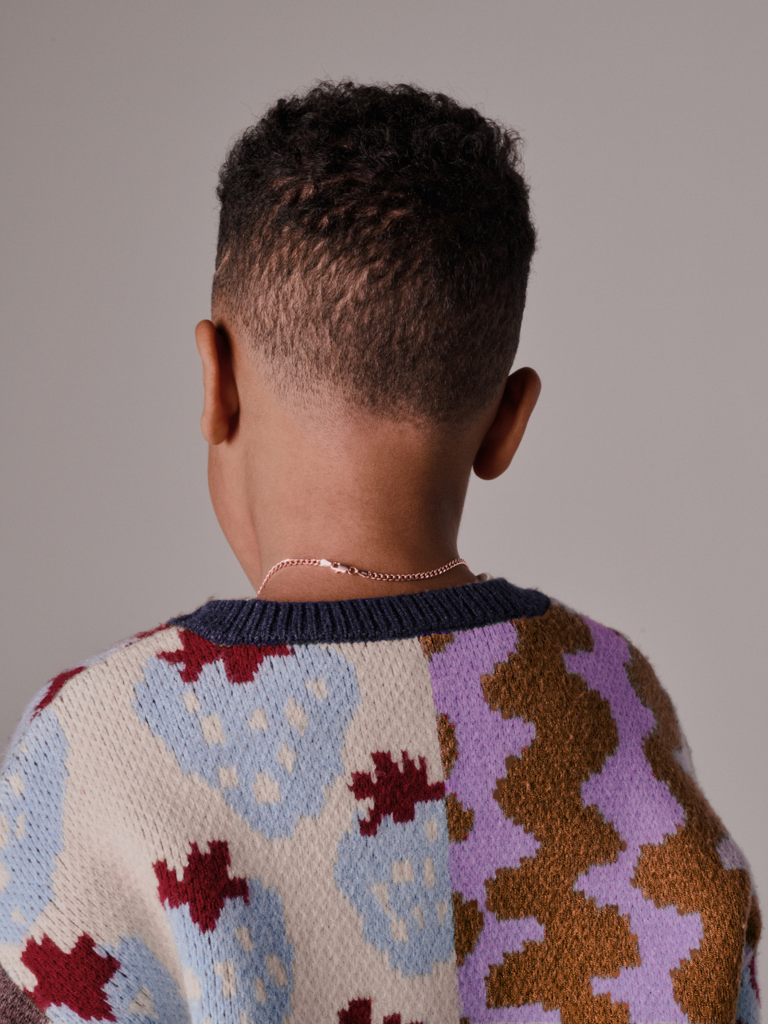 A child with short curly hair is seen from behind, wearing the EVERYTHING SWEATER featuring soft knit fabric with wavy checkers, paired with a thin chain necklace against a plain background.