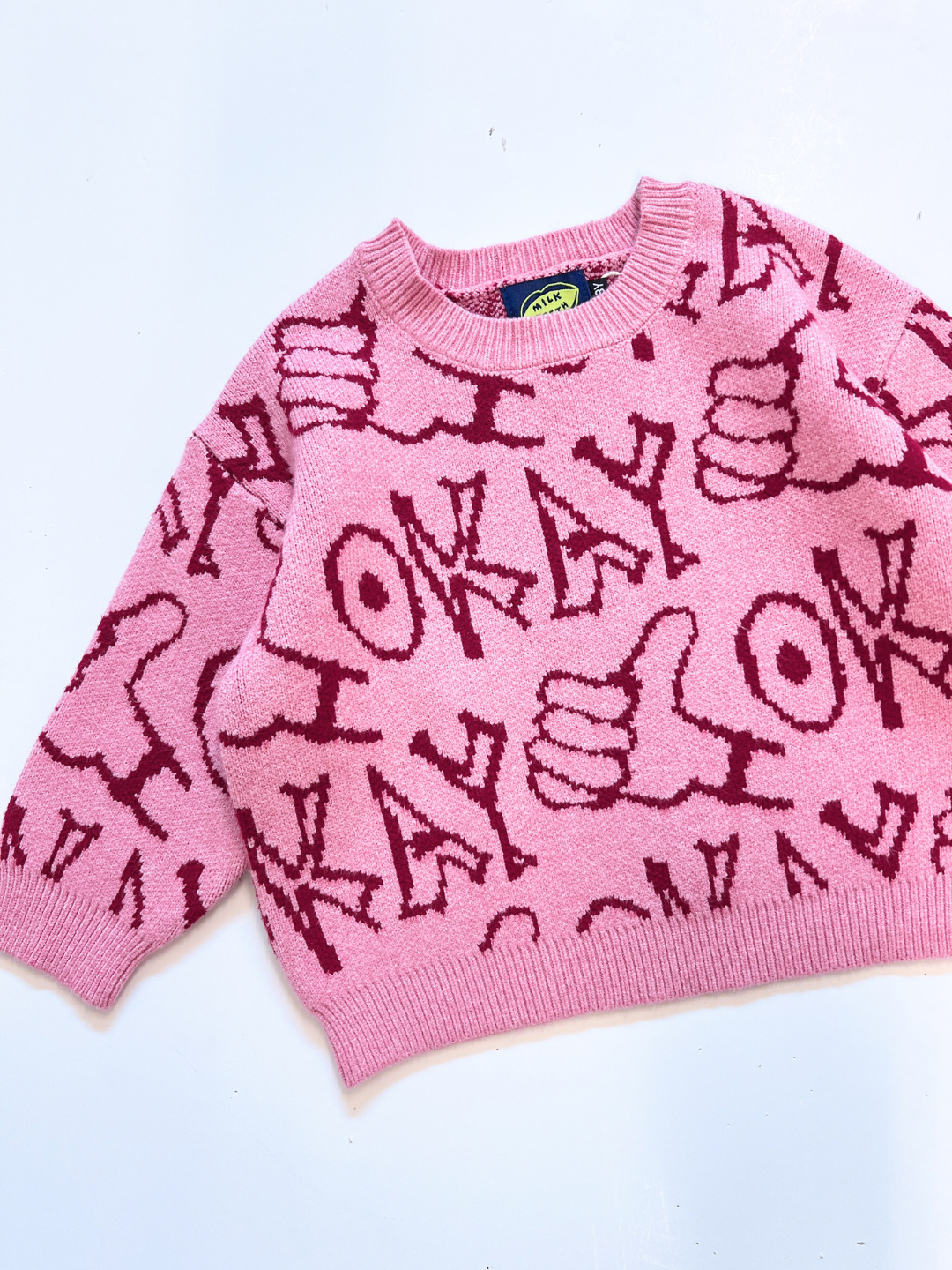 The OKAY SWEATER is a pink sweater with red patterns, showcasing the word "OKAY" and hand signs making a thumbs-up gesture.