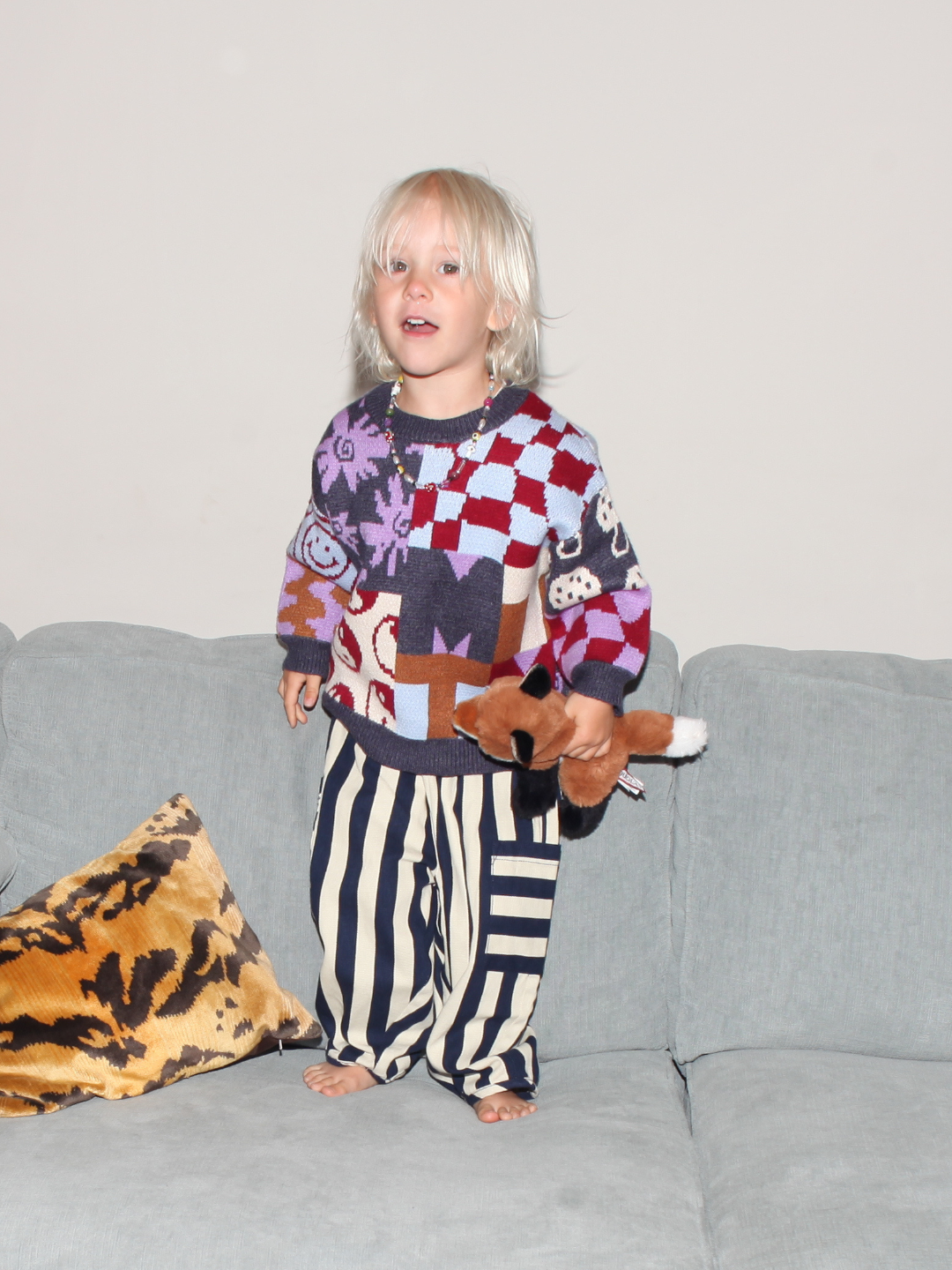 Child wearing a colorful sweater and striped pants holding a teddy bear on a couch.