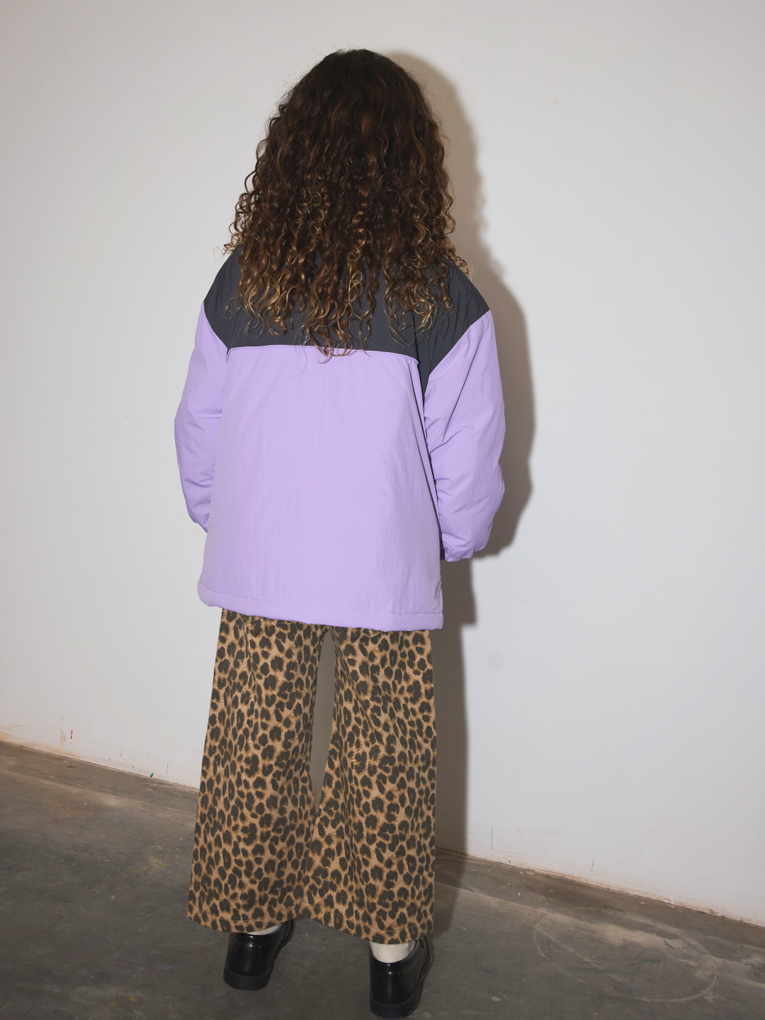 A child viewed from behind wearing a lavender and dark gray color-blocked jacket with wide-leg leopard-print pants, standing against a plain off-white wall.