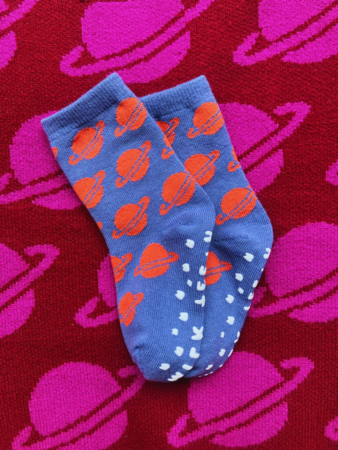 Slate Spacecamp | GRIP SOCKS, featuring a stretchy blue material adorned with orange planet patterns and white dots, are showcased on a red fabric with pink planet designs.