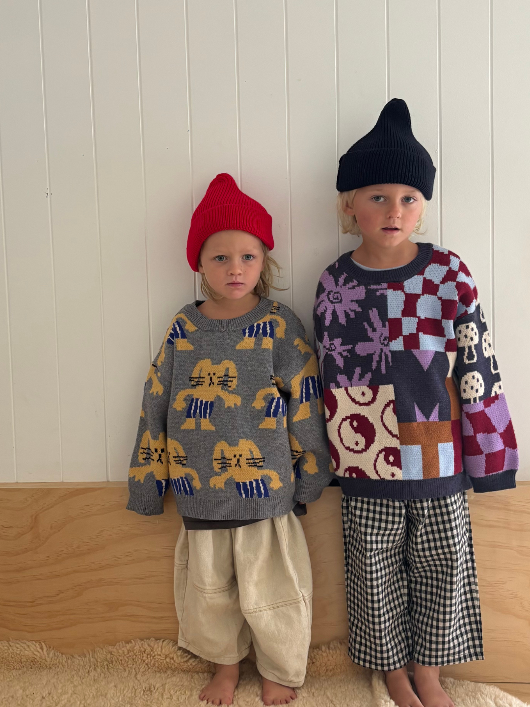 Two children wearing patterned sweaters and hats against a wooden floor and white wall background.