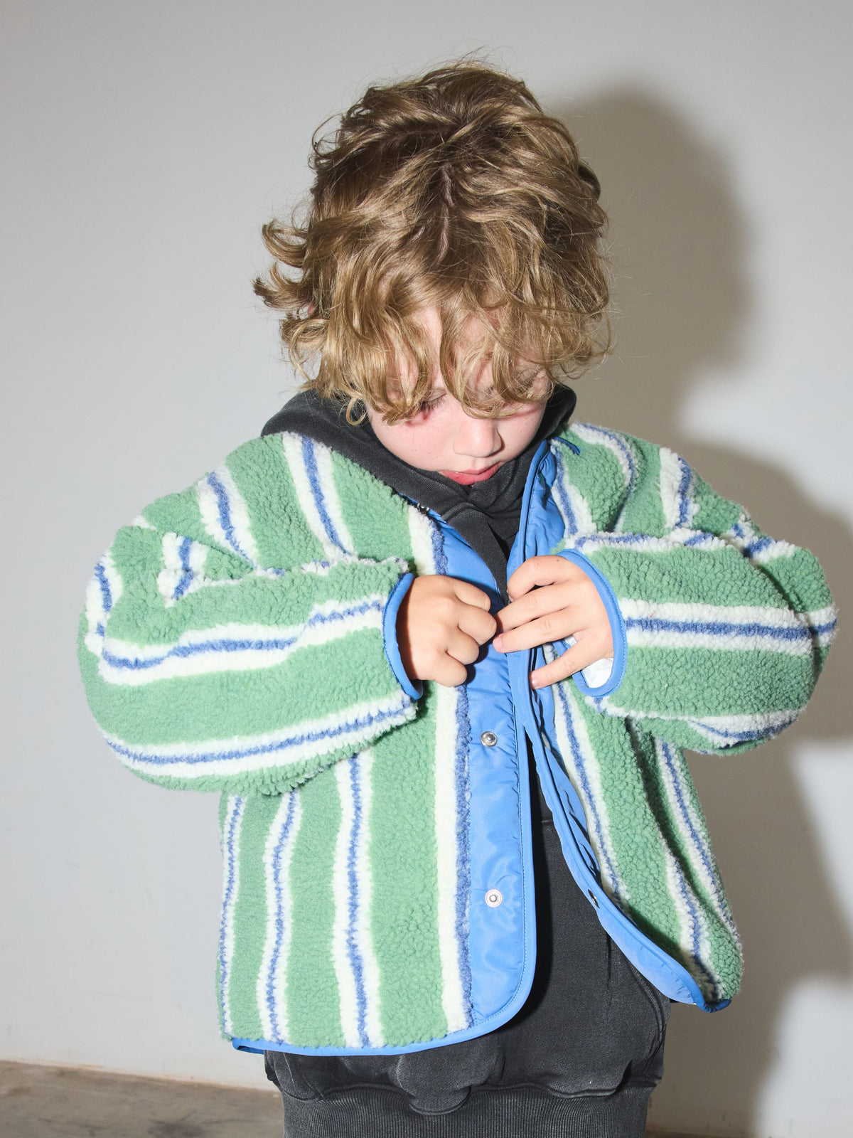 Child looking down while snapping the green striped sherpa jacket closed over a charcoal hoodie.