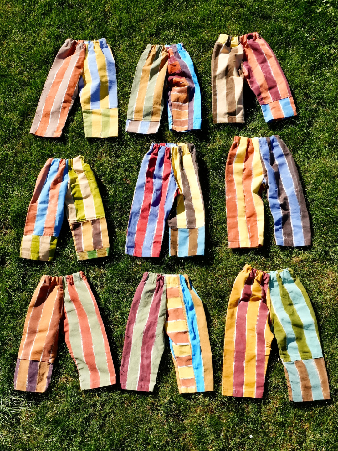 Nine pairs of PAINTER PANTS 2-3Y, each featuring vibrant, upcycled vertical stripes in unique color combinations, are laid out in three rows on green grass.