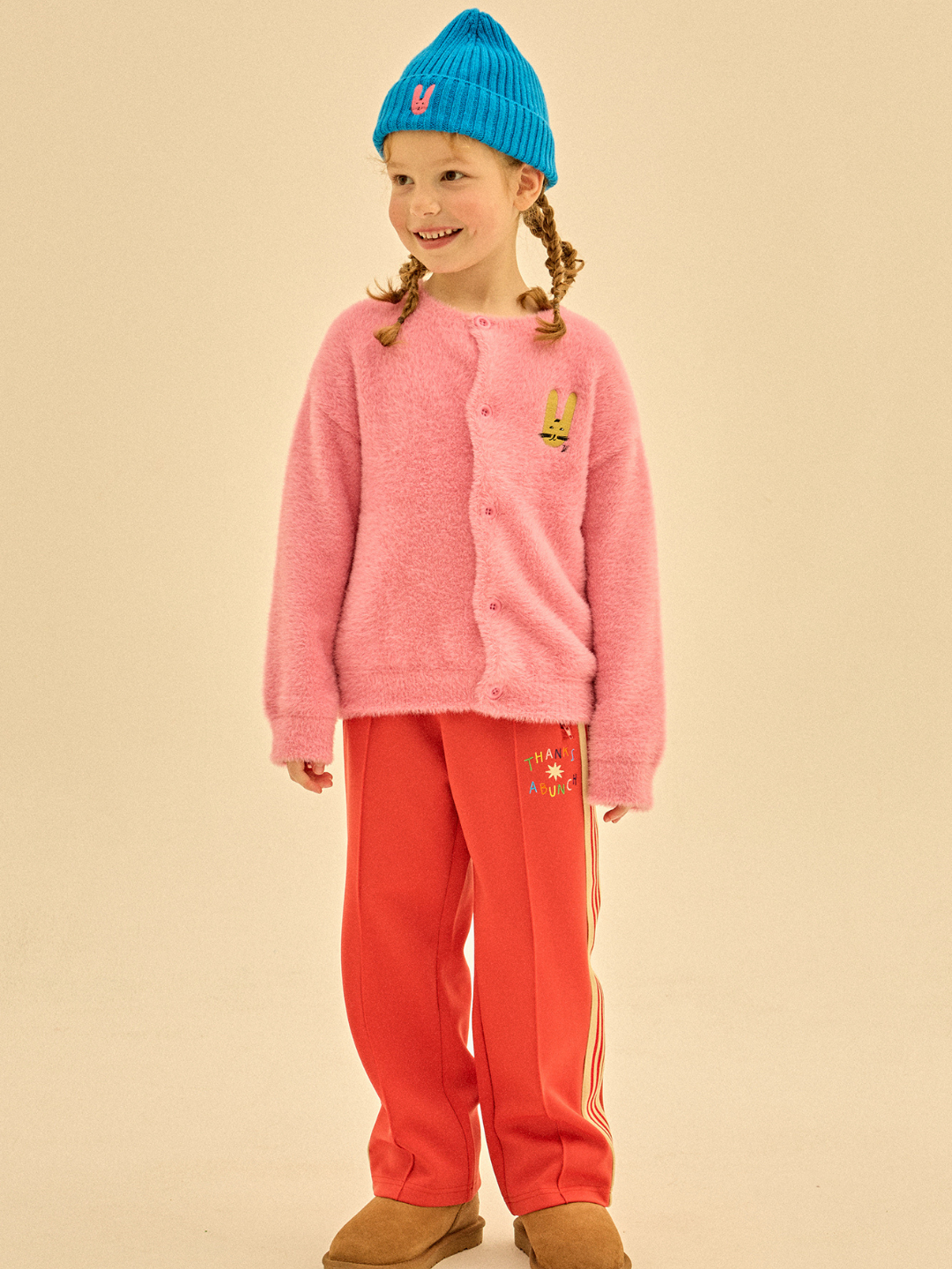 A young girl stands and smiles against a plain background, wearing a blue beanie, pink cardigan, THANKS TRACK PANTS in red, and brown shoes, beautifully showcasing the importance of careful merchandising.