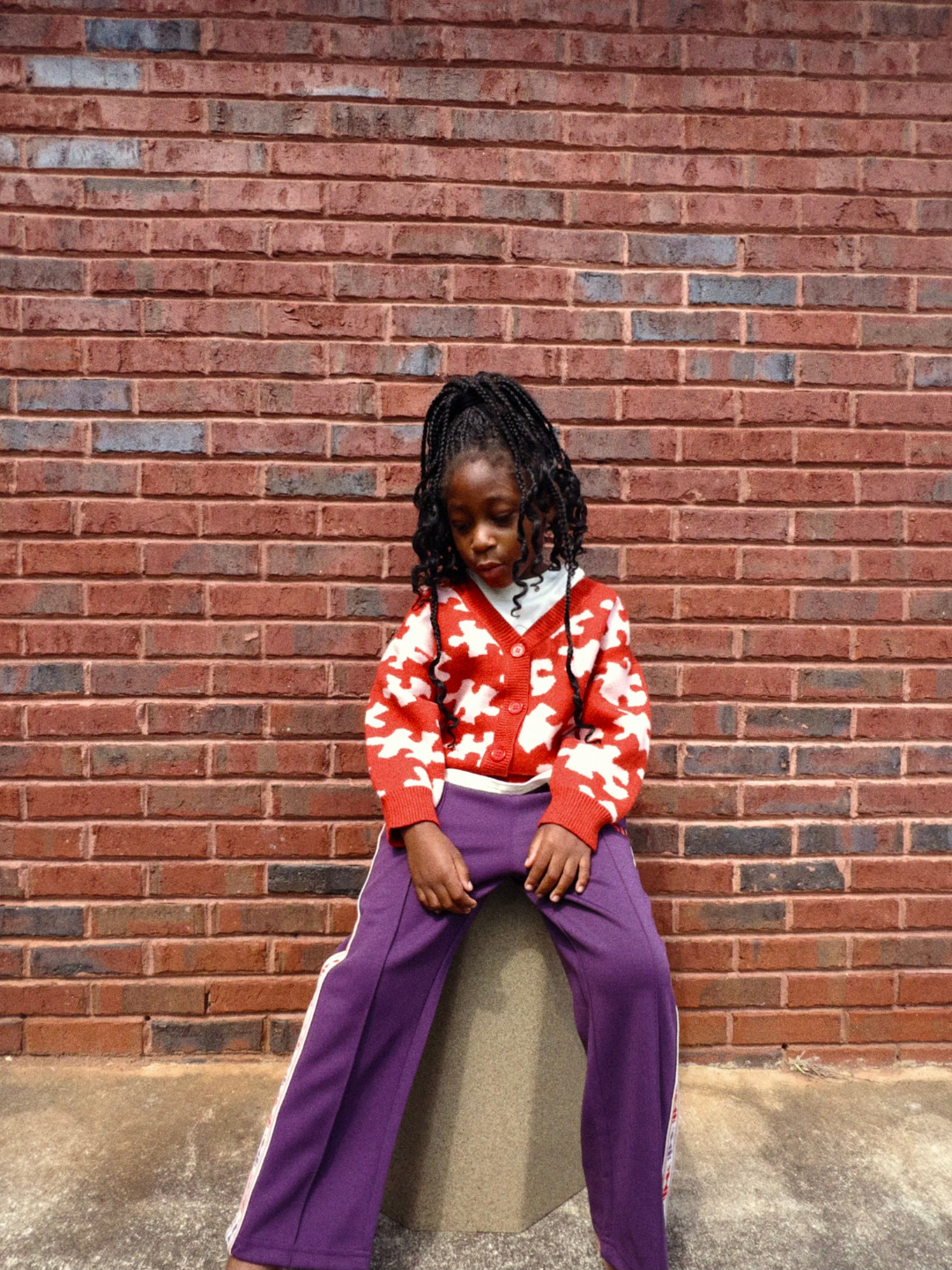 Red | Child wearing a red and white patterned sweater and purple pants sitting against a brick wall.